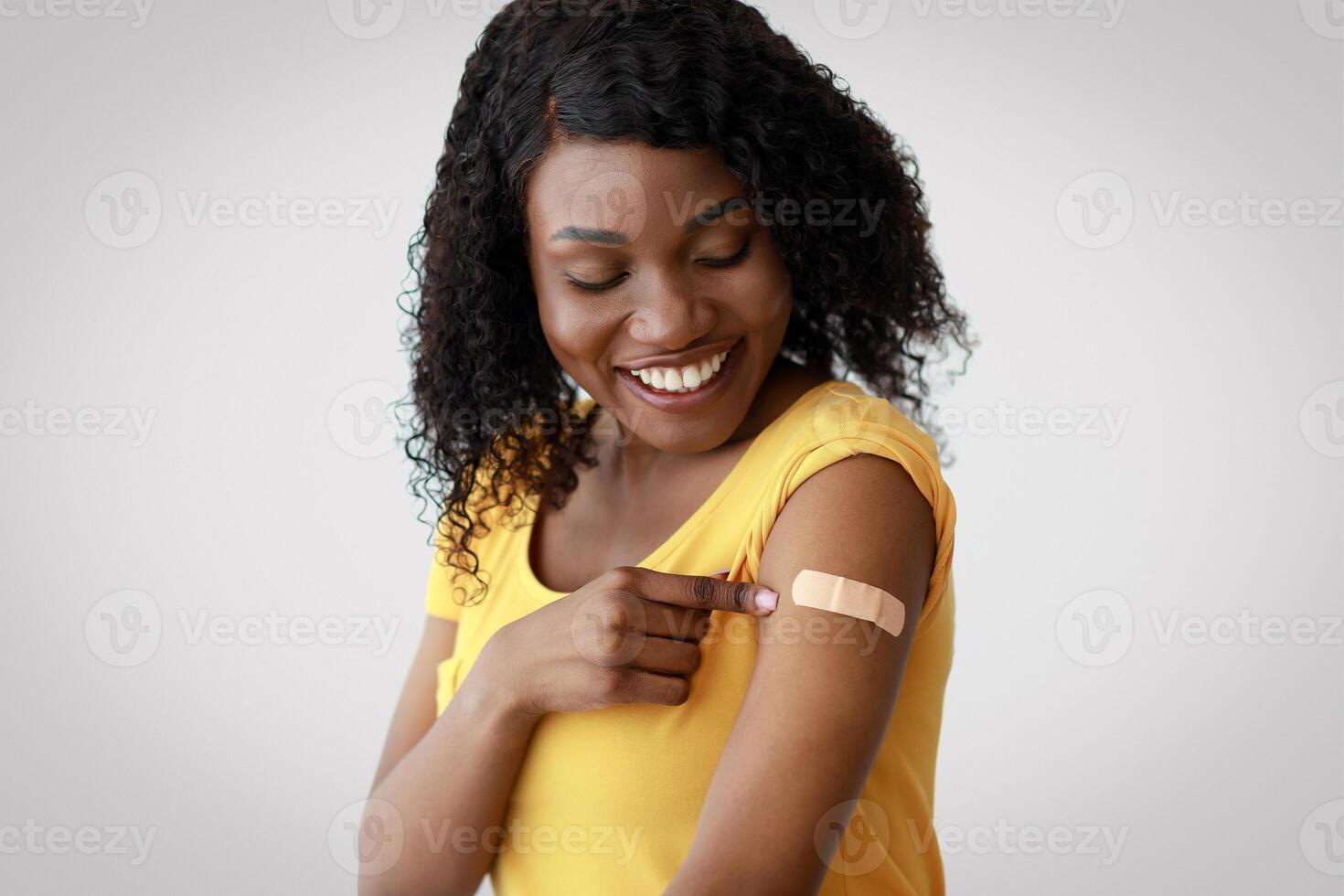 Covid-19 Vaccination. Vaccinated African American Female Looking At Arm With Plaster Bandage After Flu Antiviral Vaccine Injection Sitting Over Gray Background In Studio. Coronavirus Protection photo