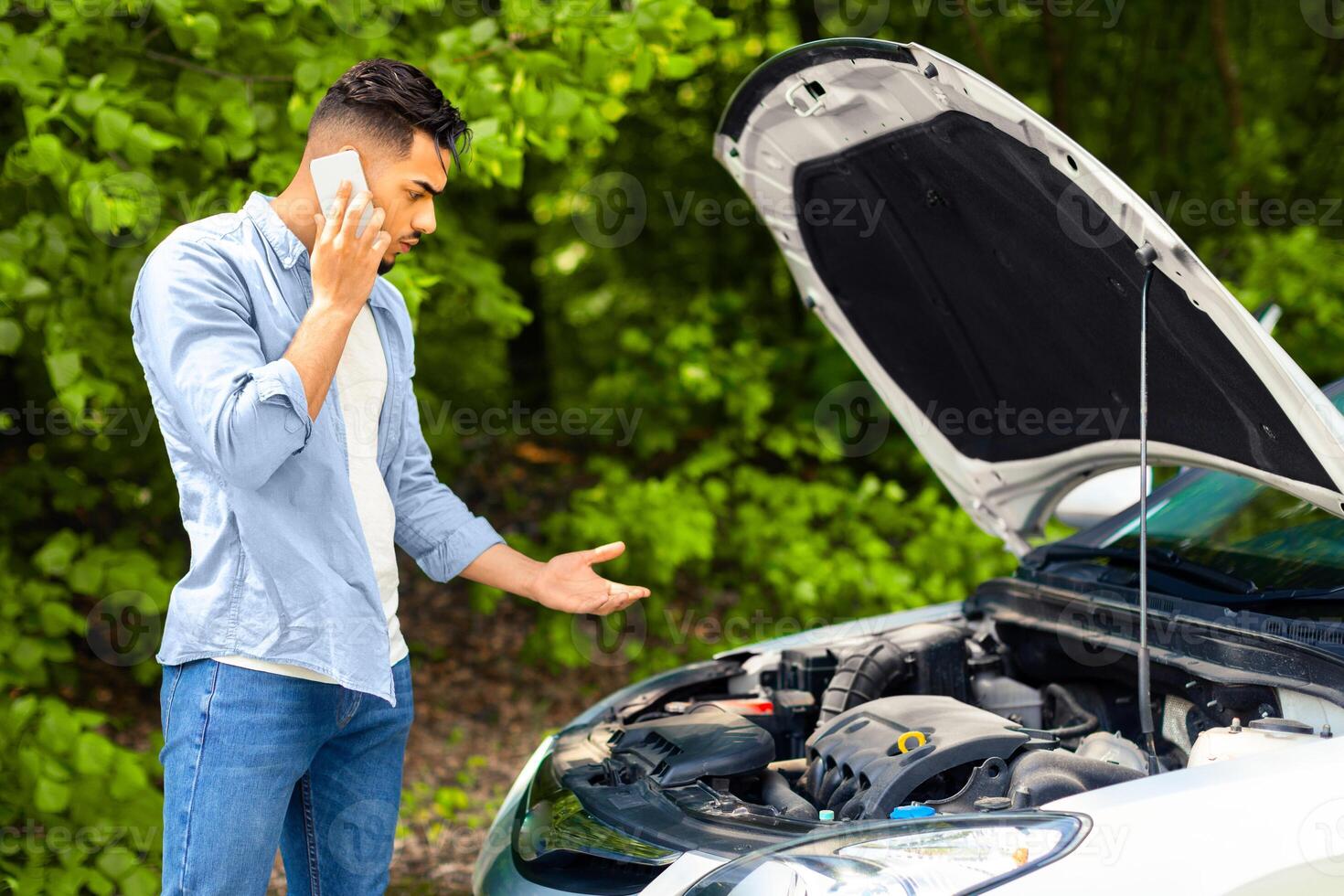 Confused arab guy driver looking under car hood, calling auto service, upset middle-eastern young man got stuck at highway, having phone conversation with friends, asking for help, side view photo