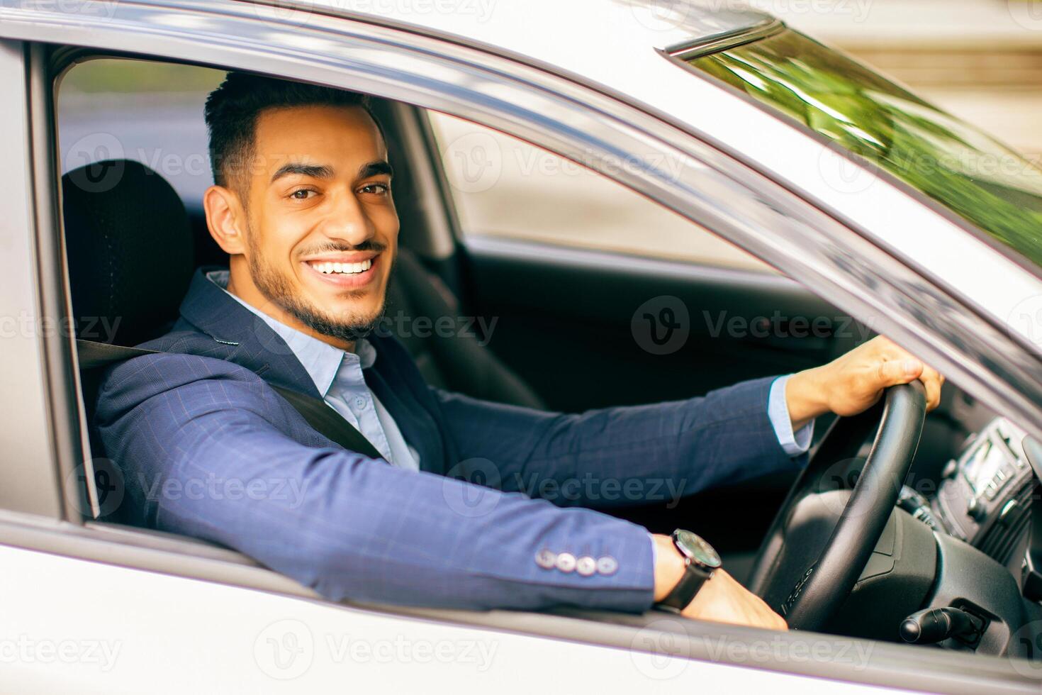 Photo of wealthy middle-eastern businessman in stylish suit driving nice car, going to office, having important business meeting, looking through window and smiling at camera, side view.