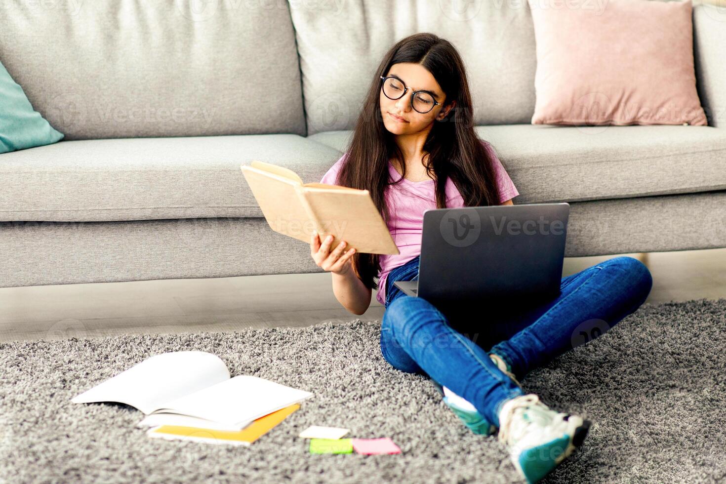 Full length of Indian teen girl reading textbook, sitting on floor with laptop and study materials, learning remotely from home, copy space. Focused adolescent getting ready for online webinar photo