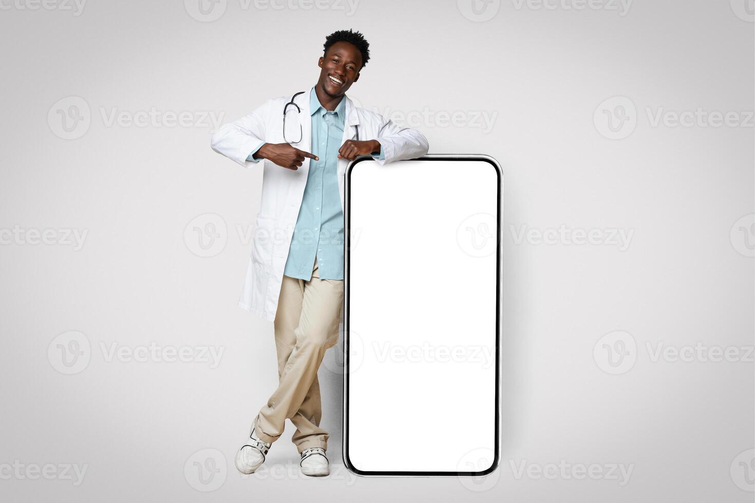 Black Young Male Doctor In Uniform Leaning And Pointing At Big Smartphone With Blank Screen, Showing Free Copy Space For Coronavirus Info Or Vaccination Campaign Design, Standing On Beige Background photo