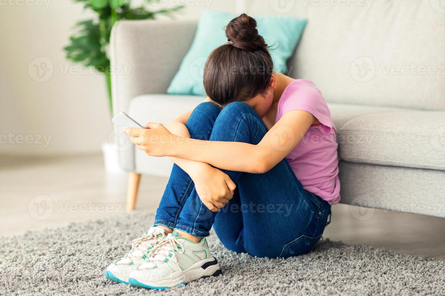Depressed Indian teen girl with cellphone crying on floor at home, suffering from cyber bullying, being harassed online. Stressed teenager being victim of internet abuse or trolling photo