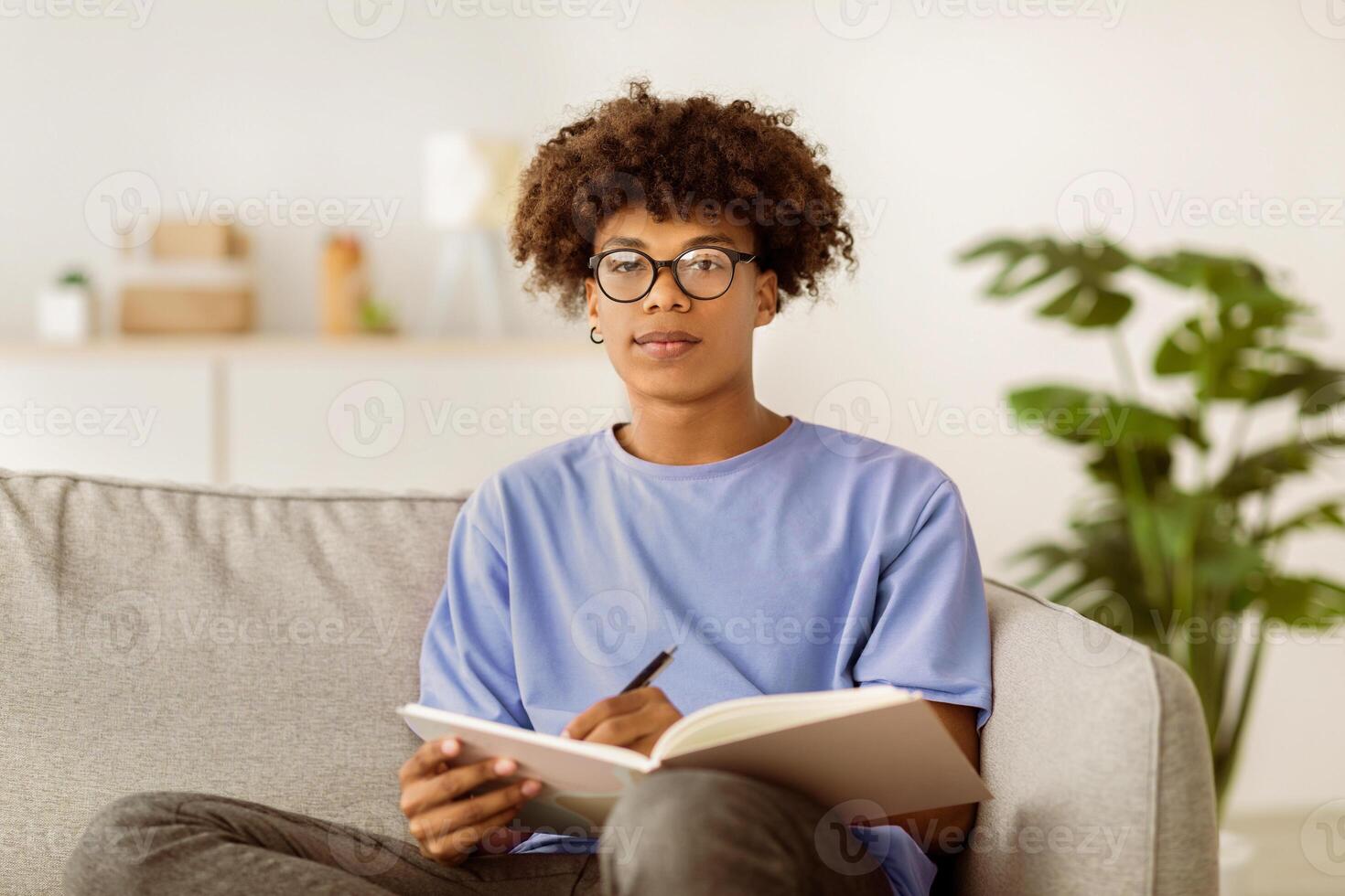 Focused black teenager writing in notebook, getting ready for exam or test, sitting on couch indoors. Cool African American adolescent studying from home during covid lockdown photo
