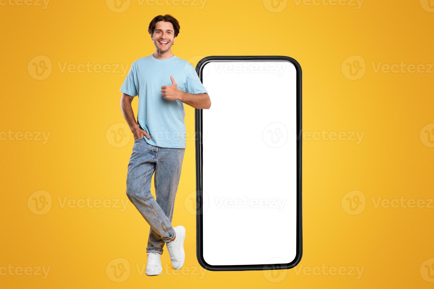 Happy Man Standing Near Big Smartphone With Empty White Display Gesturing Thumb Up Sign, Cheerful Casual Male Recommending New App Or Website, Standing On Yellow Background, Mock Up, Full Body Length photo
