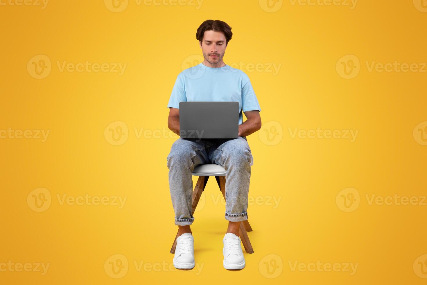 Portrait of serious bearded guy sitting on chair using laptop computer, reading news, watching or movie, working or elearning remotely, isolated on orange studio background, front vertical shot photo