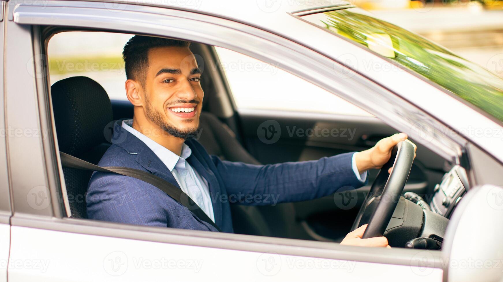 Happy middle-eastern young businessman driving new auto, going to office, shot from outside, side view. Handsome successful arab guy manager going home after good day, looking at camera and smiling photo