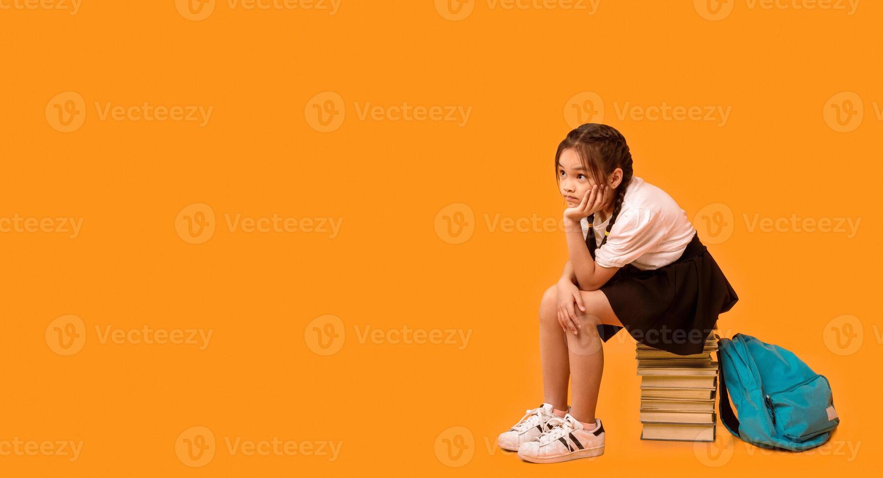 Unhappy Chinese School Girl Sitting On Book Stack Waiting For Class Over Yellow Studio Background. Free Space, Panorama photo
