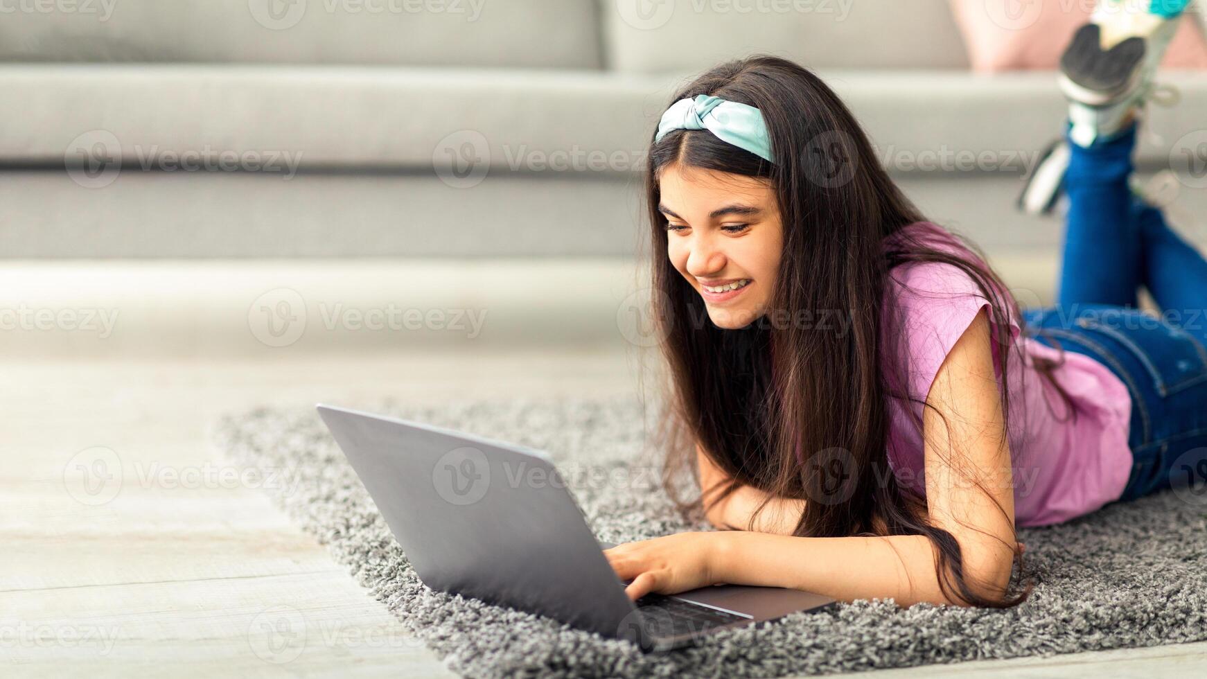 Distance learning concept. Joyful adolescent girl studying online, lying on soft carpet with laptop indoors. Indian teenager using computer, doing home assignment, communicating to friend on webcam photo