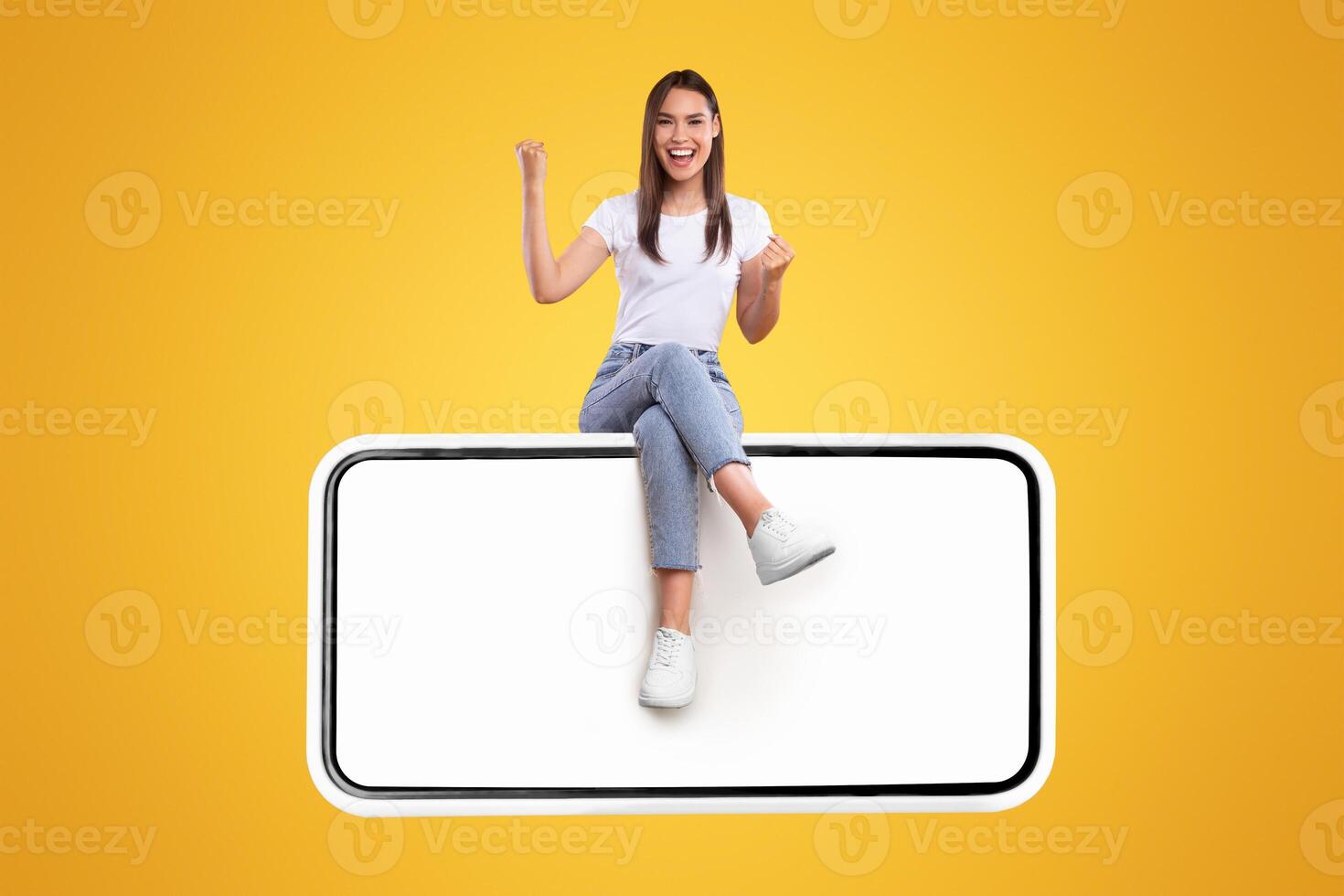 Yes. Excited Woman Sitting On Big Smartphone With Empty White Screen Shaking Clenched Fists, Cheerful Lady Celebrating Win Isolated On Yellow Orange Background, Mock Up Collage, Full Body Length photo