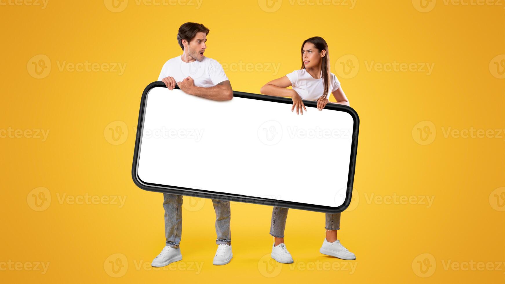 It's Mine. Couple holding big smartphone with white empty screen on yellow orange studio background fighting for gadget pulling device in different directions. Mock up display template, banner photo