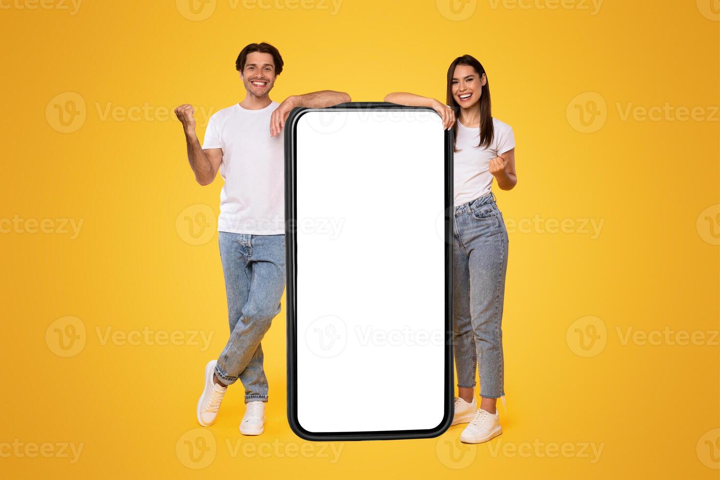 Yes. Excited Couple Leaning On Big Smartphone With Empty White Screen Shaking Clenched Fists, Cheerful Guy And Lady Celebrating Win, Standing On Yellow Background, Mock Up Collage, Full Body Length photo