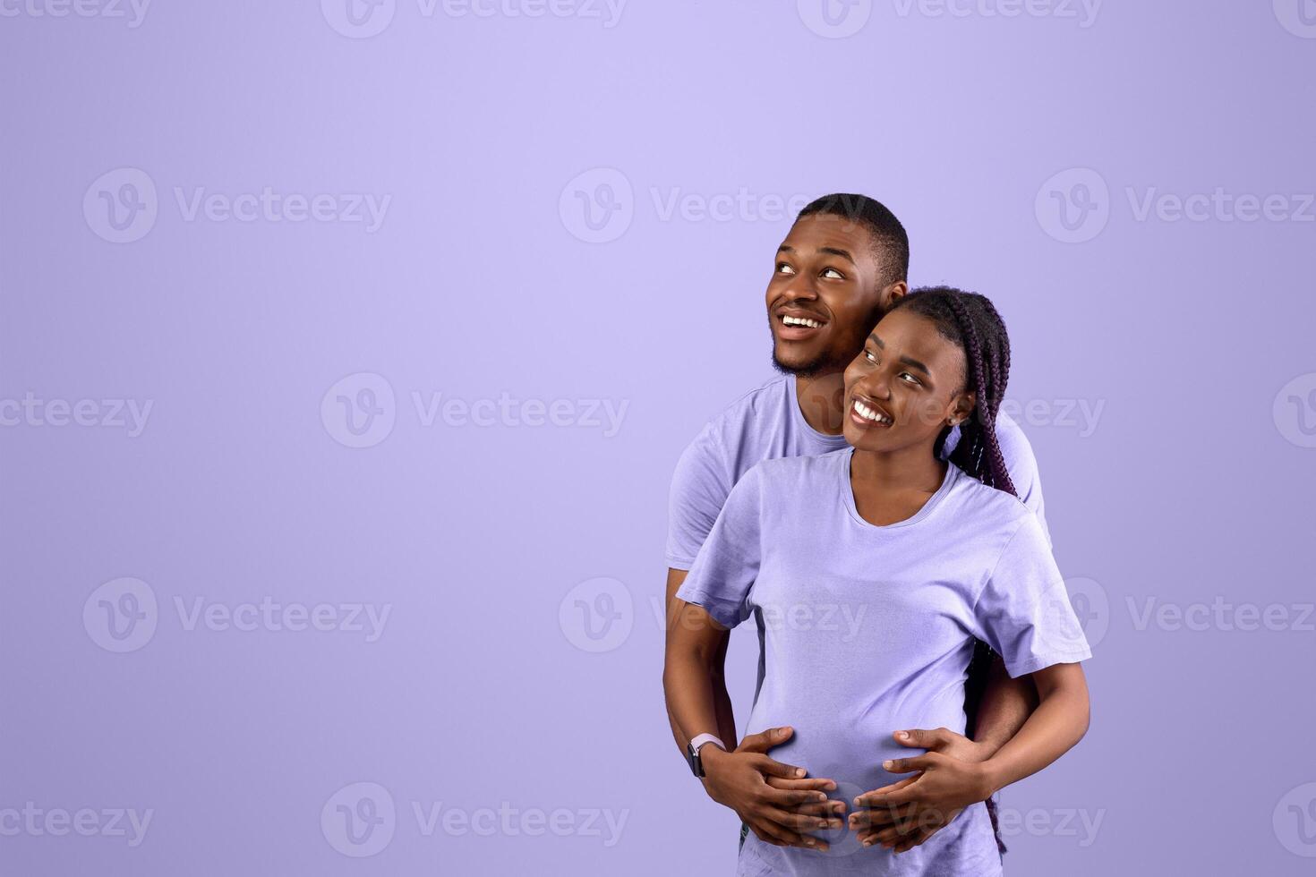Smiling black husband hugging pregnant wife from back, cheerful couple standing on purple violet studio background, thinking about baby's name or gender looking up at empty speech bubble, free space photo