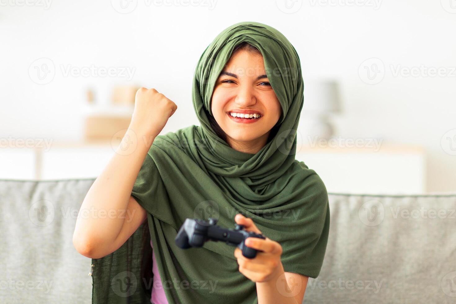 Triumphant Indian teen girl in hijab with joystick playing videogame, making YES gesture, celebrating victory at home. Positive adolescent in headscarf engaged in computer game photo
