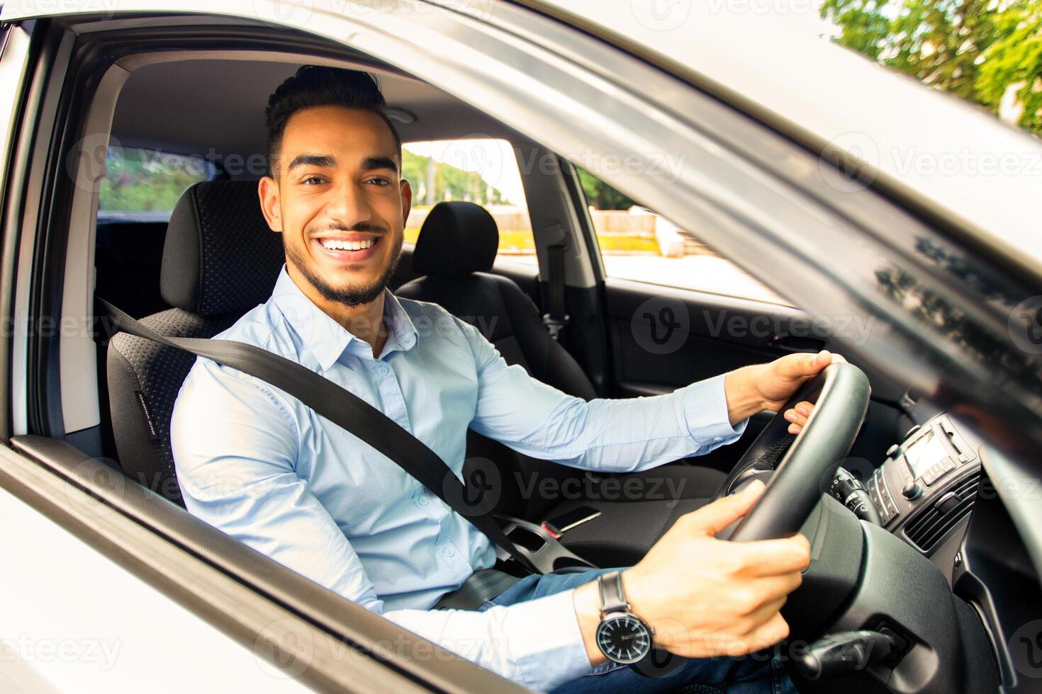 Cheerful arab businessman driving car, shot from outside, going to office or business trip, copy space. Happy middle-eastern guy in formal outfit with fasten seat belt driving his brand new car photo