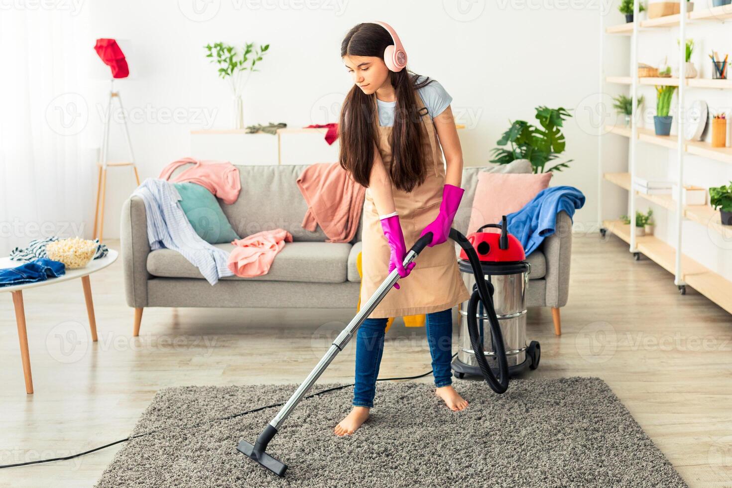 Full length of Indian teen girl cleaning her apartment, wearing wireless headphones, listening to music while vacuuming floor. Cool adolescent doing house chores, performing domestic cleanup photo