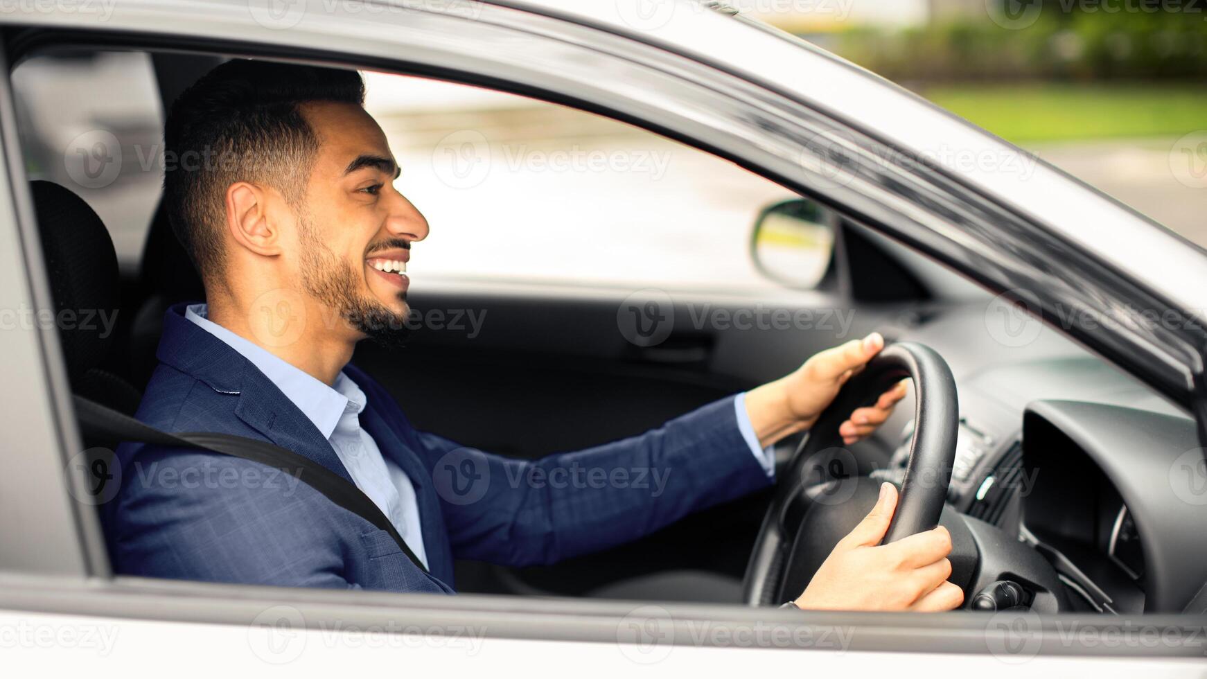 Happy middle eastern young businessman driving new auto, going to office, shot from outside, side view. Handsome successful arab guy manager going home after good day, looking at road and smiling photo