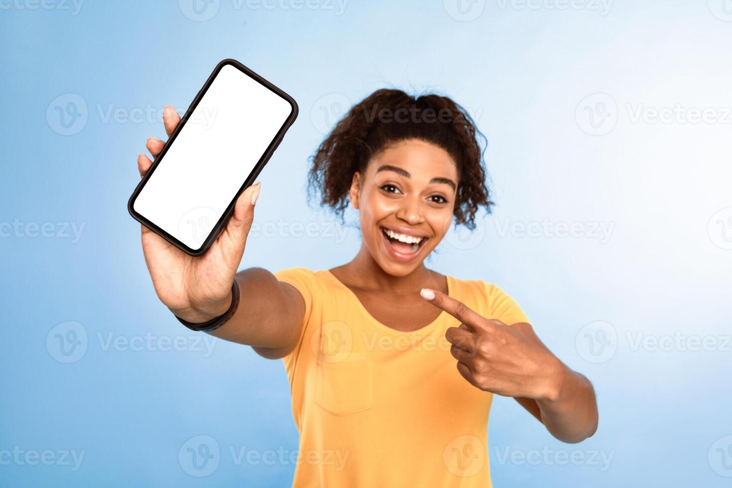 Check this out, cellphone display mockup. Excited African American woman pointing at smartphone with empty screen on blue background, selective focus. Space for mobile app, website or online ad photo