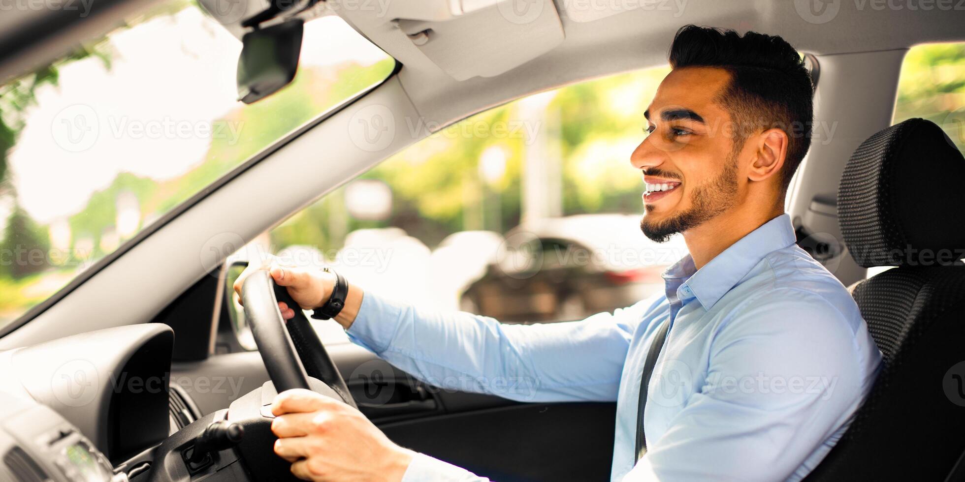 Joyful bearded arabic guy in formal outfit looking at road and smiling, driving nice automobile, young middle eastern businessman going to office, side view, copy space, sun flare photo