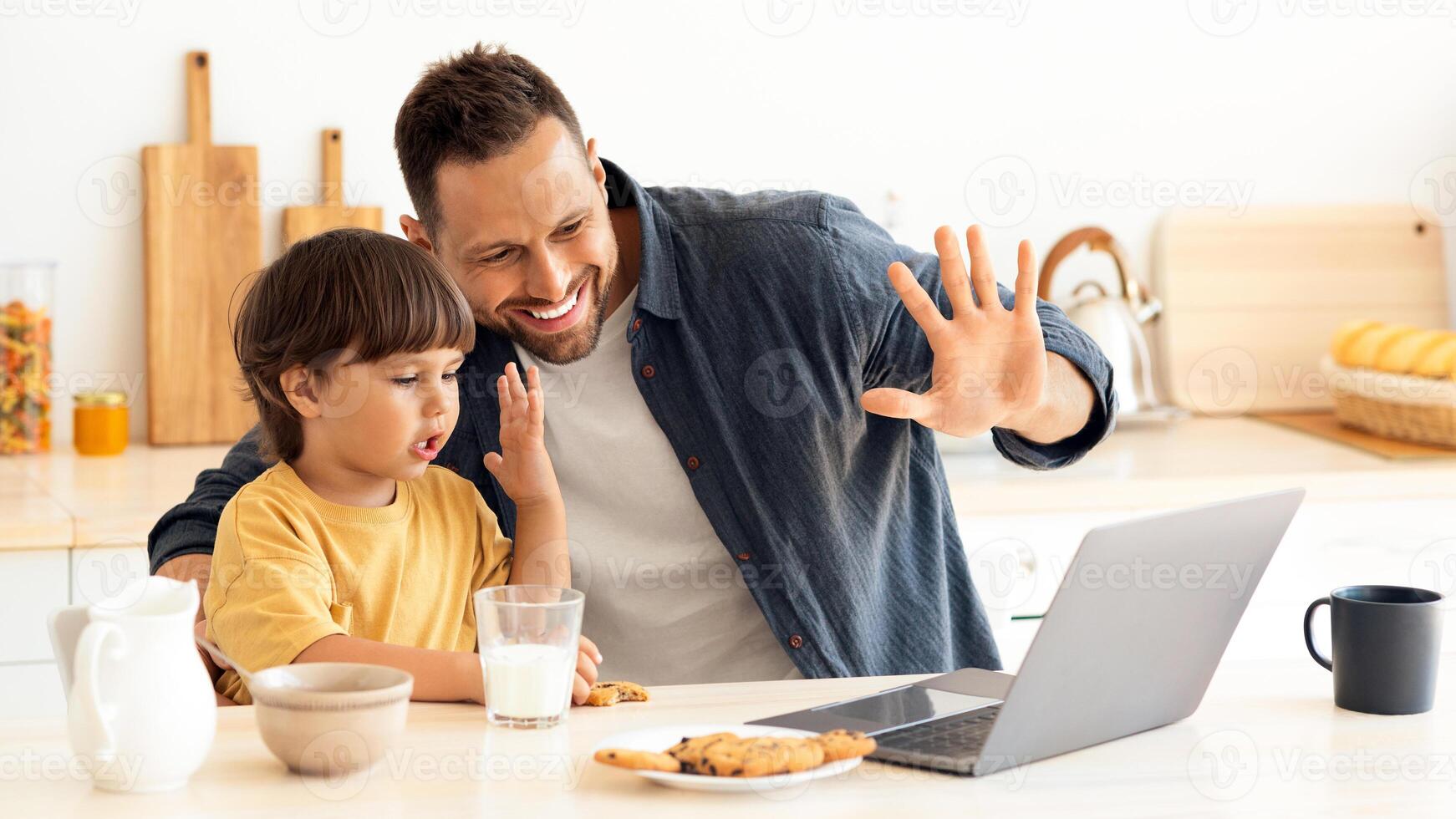 Online communication. Little boy and his daddy calling to relatives via laptop, waving hands to computer and smiling, sitting together at kitchen, free space photo