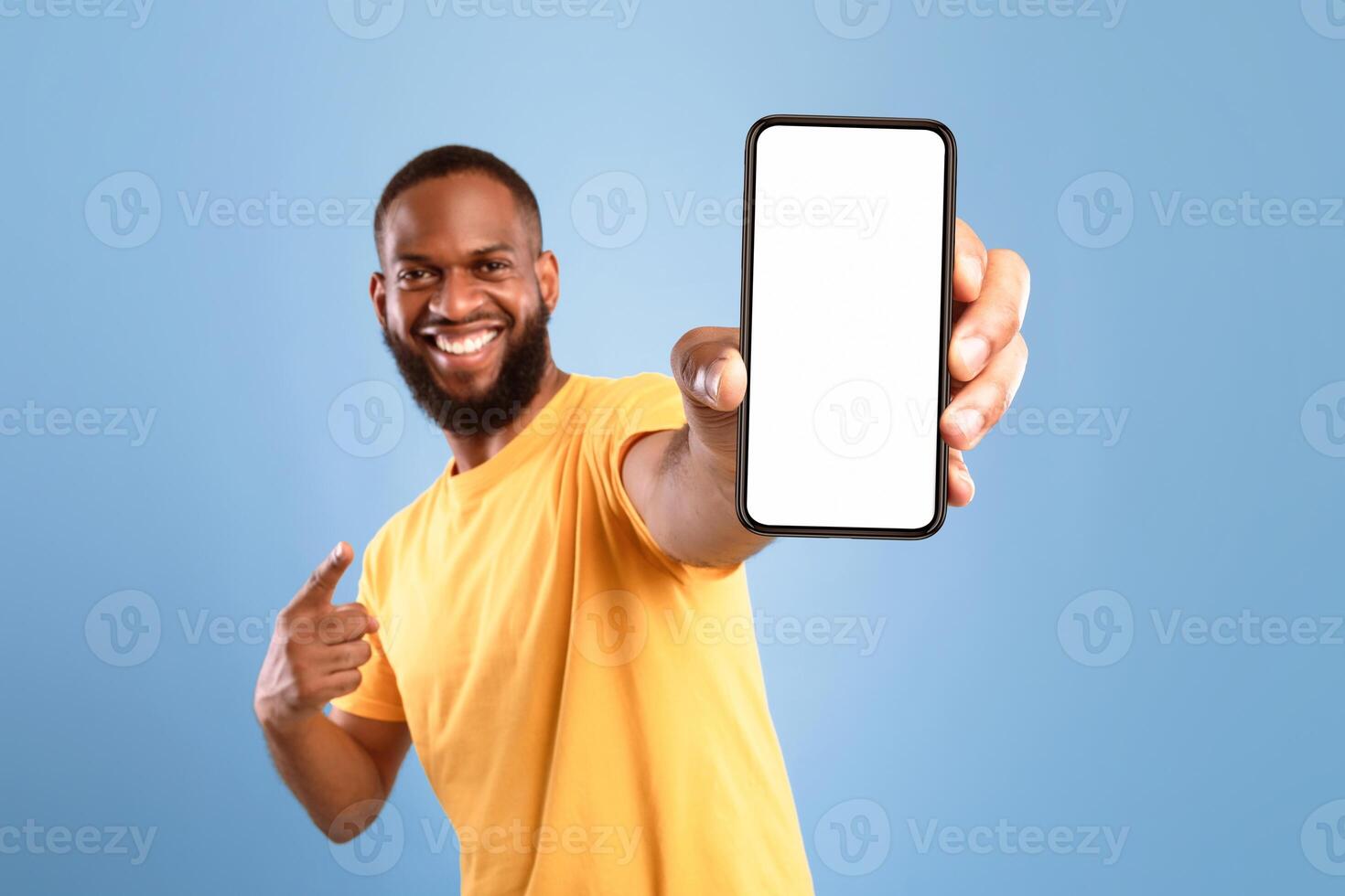 Excited African American guy in casual wear pointing at cellphone with empty screen on blue studio background, space for website or mobile app design. Cellphone display mockup. Selective focus photo