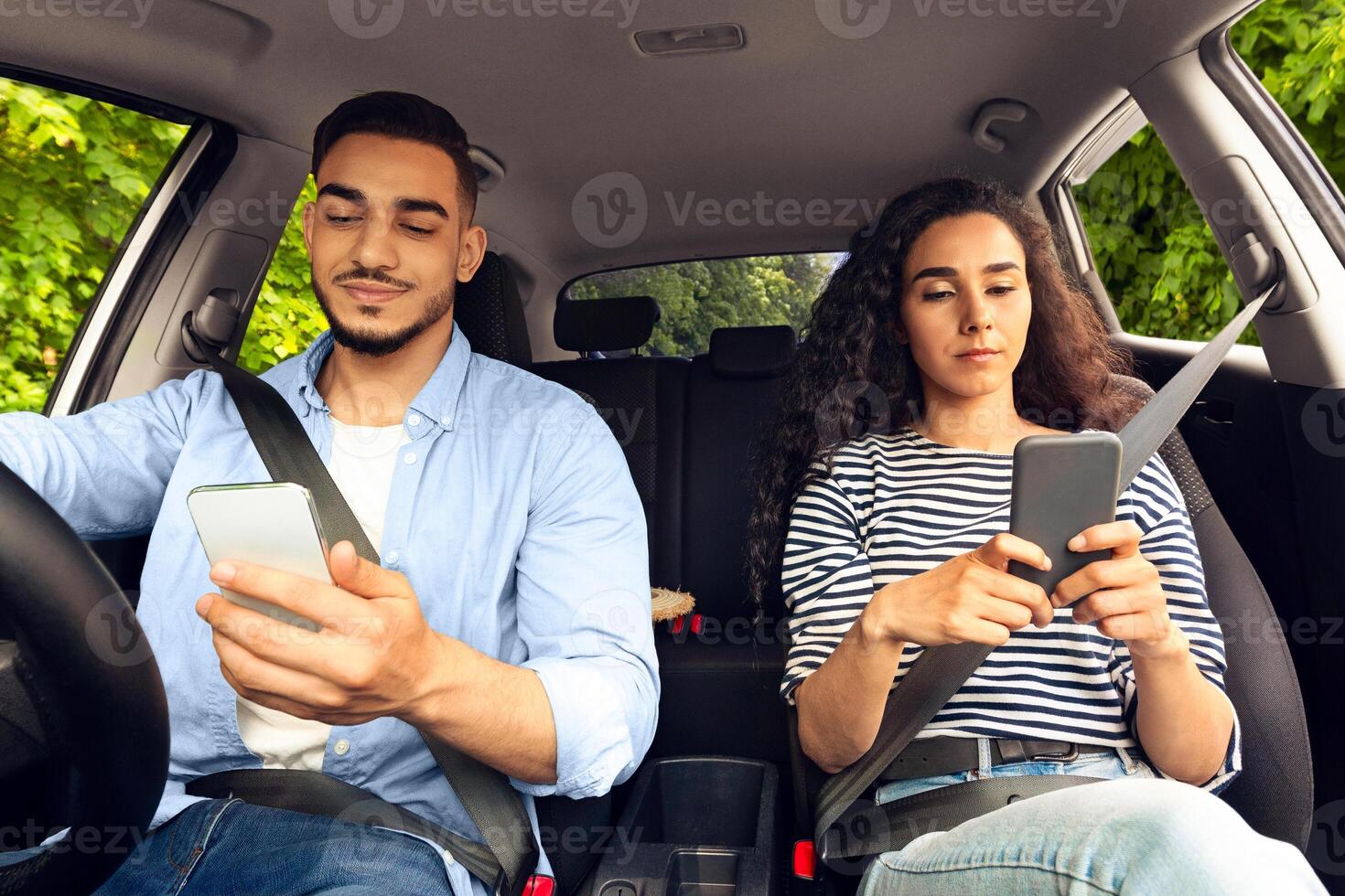Defensive, not safety driving concept. Young arabic man and woman using smartphones while travelling by car together, gadget addicted millennials sitting inside nice auto, checking social media photo
