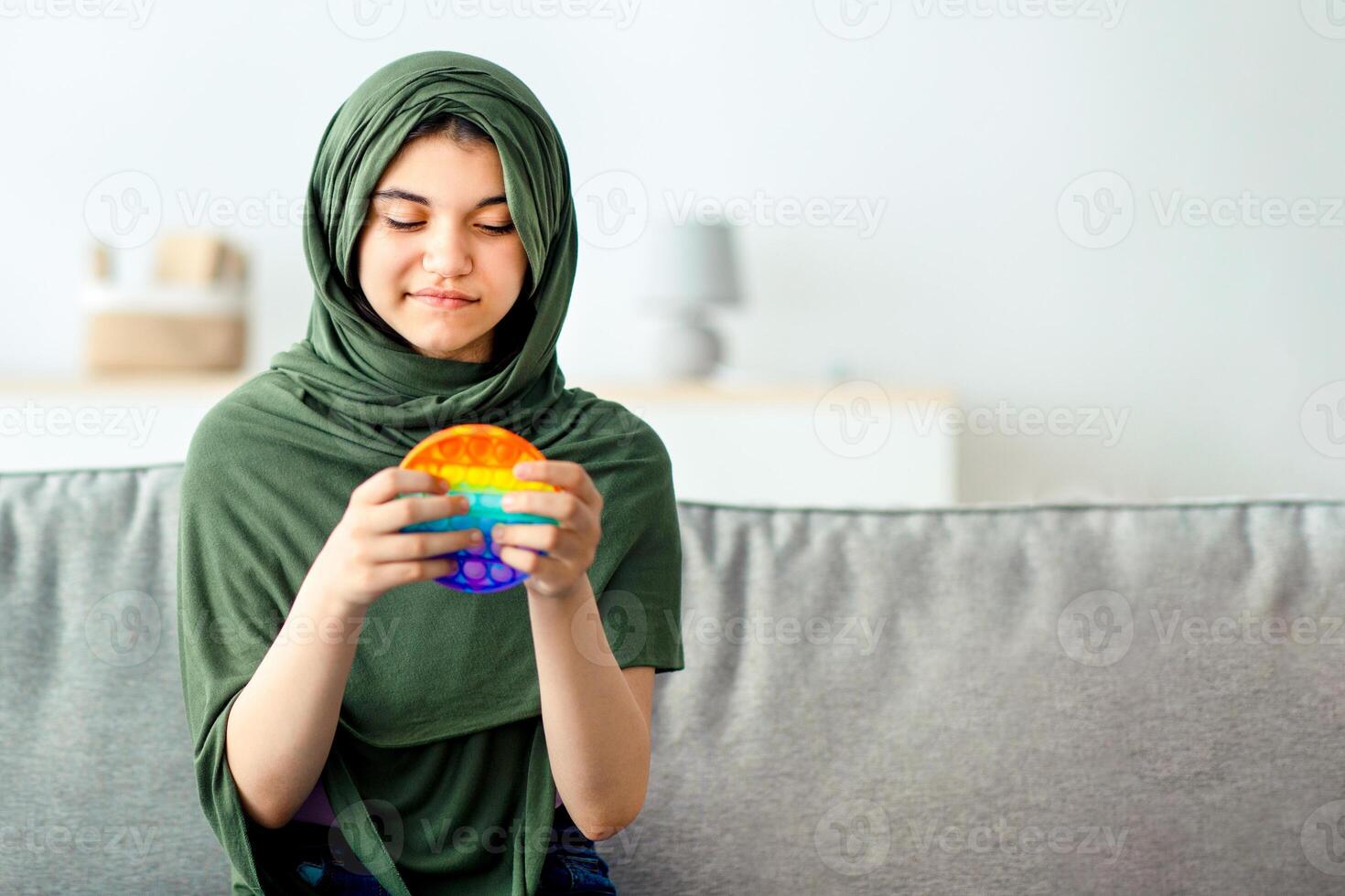 Portrait of lovely Indian teen girl in hijab playing with bright POP IT toy, enjoying fun antistress game at home. Muslim adolescent in traditional headscarf trying out modern popular plaything photo