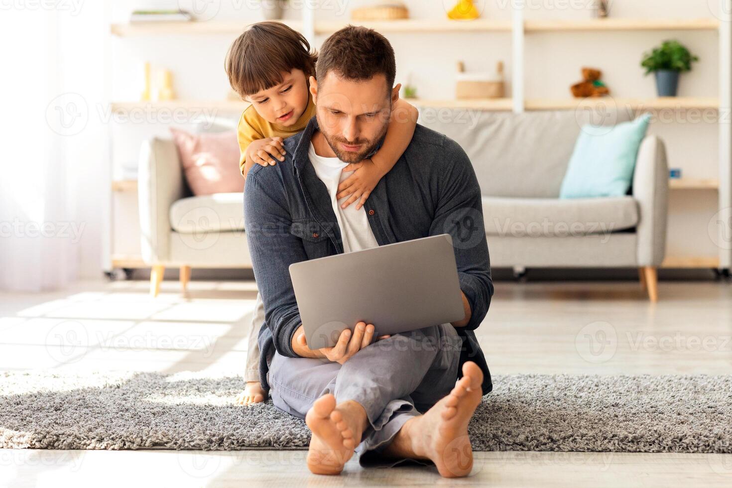 Interest for modern technologies. Young busy man working on laptop from home, his cute little son hugging him and watching at gadget, sitting together on floor at home, free space photo