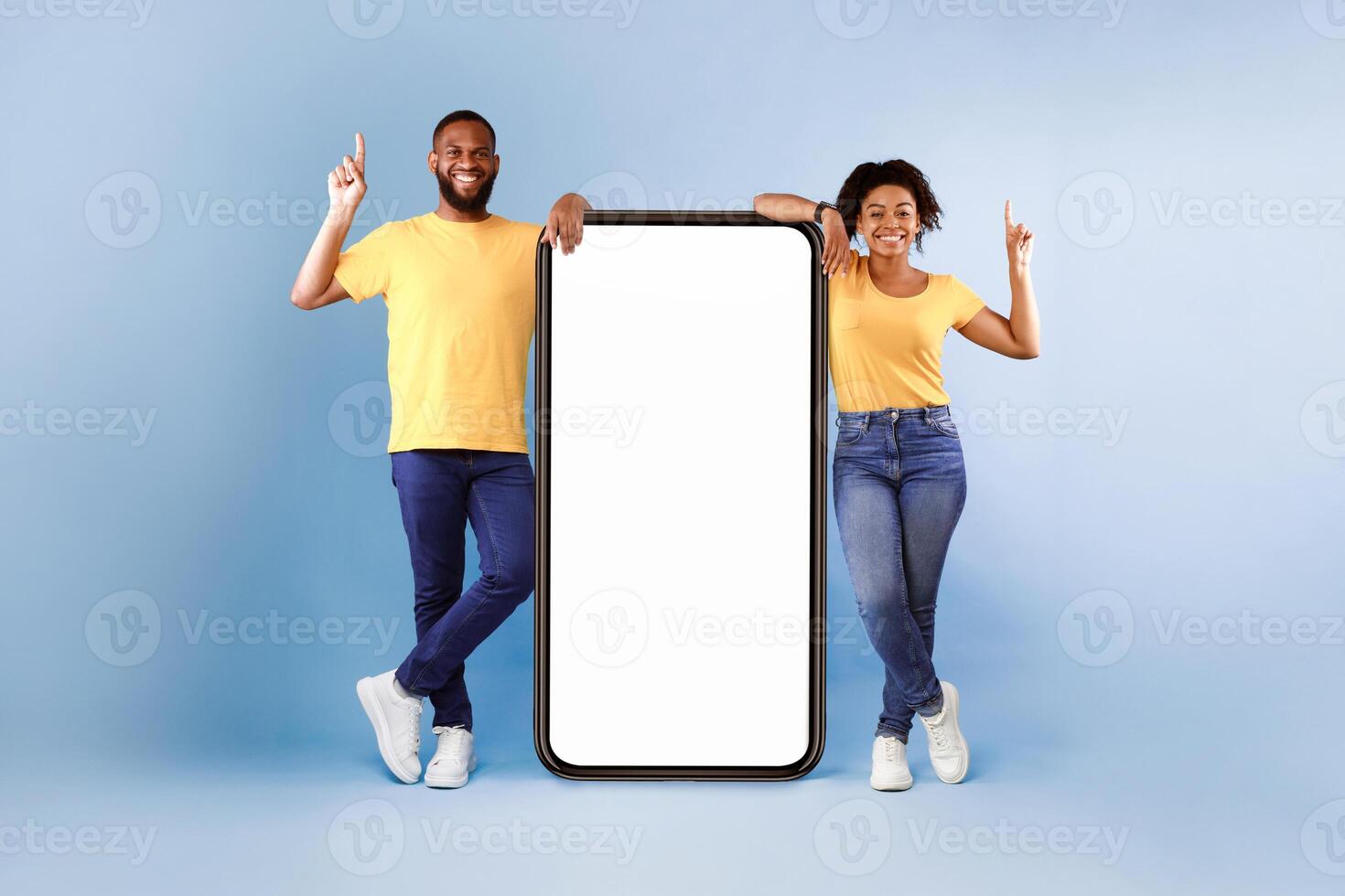 Cheerful African American couple standing next to big smartphone with blank white screen, pointing upwards, recommending new mobile application on blue studio background photo