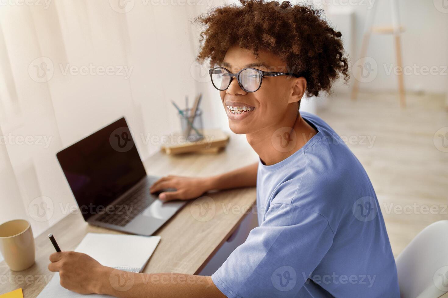 Happy black adolescent sitting at desk with laptop, learning remotely, taking notes in copybook at home, copy space. Cheerful African American teen student studying online during covid quarantine photo