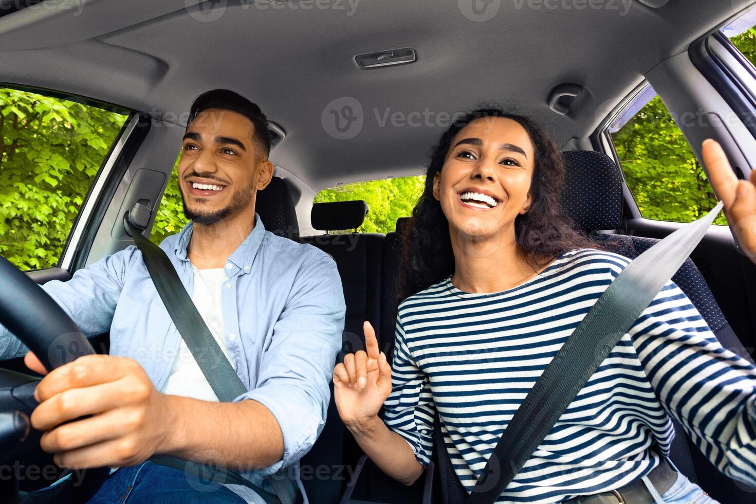 Emotional millennial mixed race couple handsome arab guy and pretty brunette lady having fun while car trip, listening to music and cherfully singing, shot from inside their automobile photo