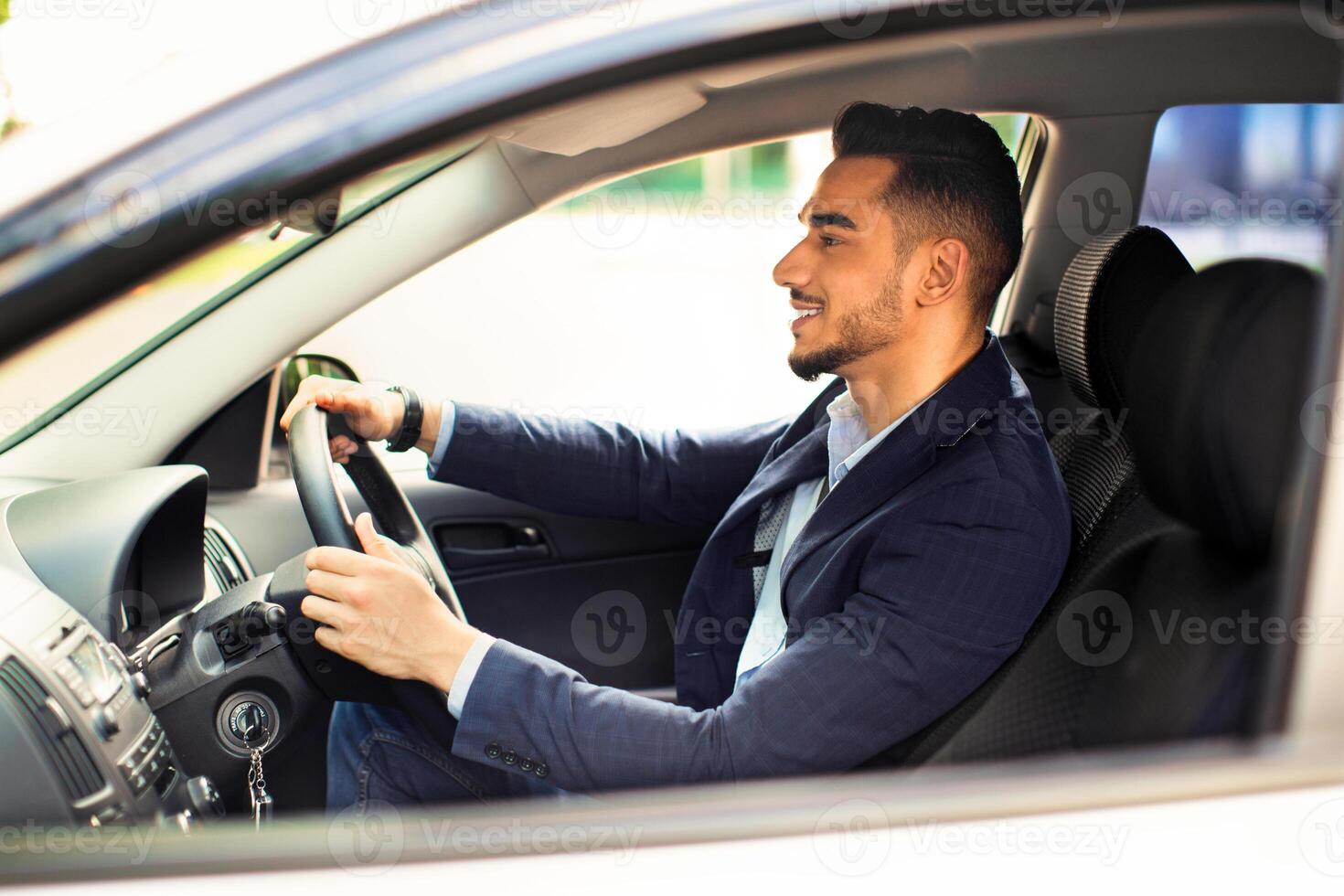 Cheerful middle eastern young man in suit manager driving white car, happy arabic businessman going to office, business meeting, side view, shot from outside, copy space, panorama photo