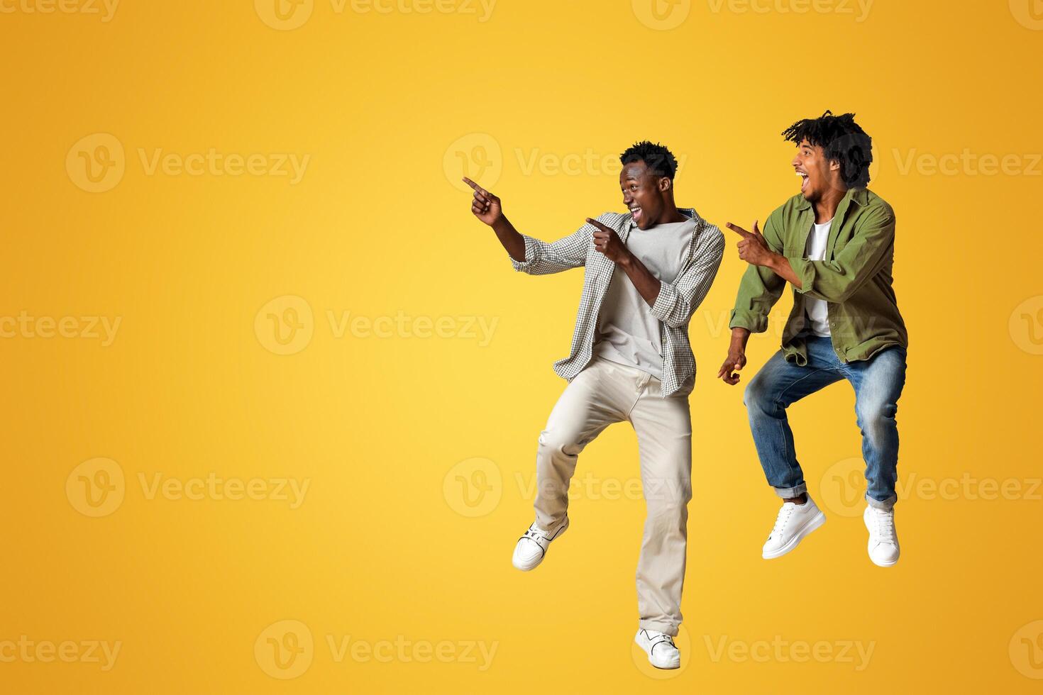 Look At This. Two Joyful Black Guys Pointing Aside While Jumping In Air Over Beige Studio Background, Cheerful African American Friends Having Fun And Demonstrating Free Copy Space, Panorama photo