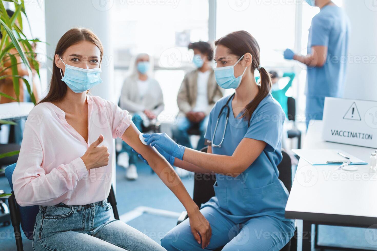 Young woman patient in protective face mask getting injection at hospital showing thumbs up gesture. Doctor cleaning disinfecting skin before giving jab rubbing arm with cotton pad at modern clinic photo