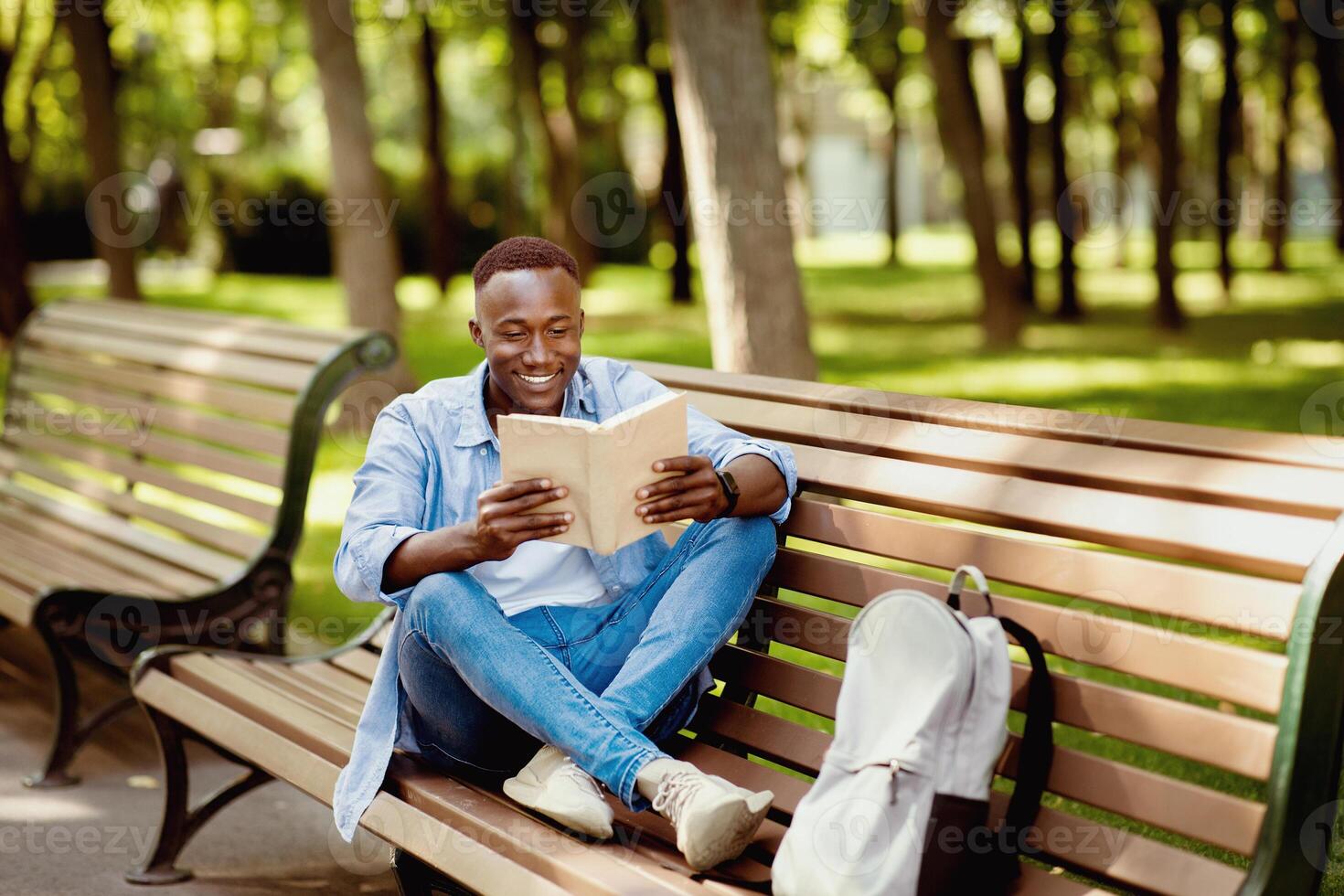 Positive black guy reading interesting book on bench at green summer park, copy space photo