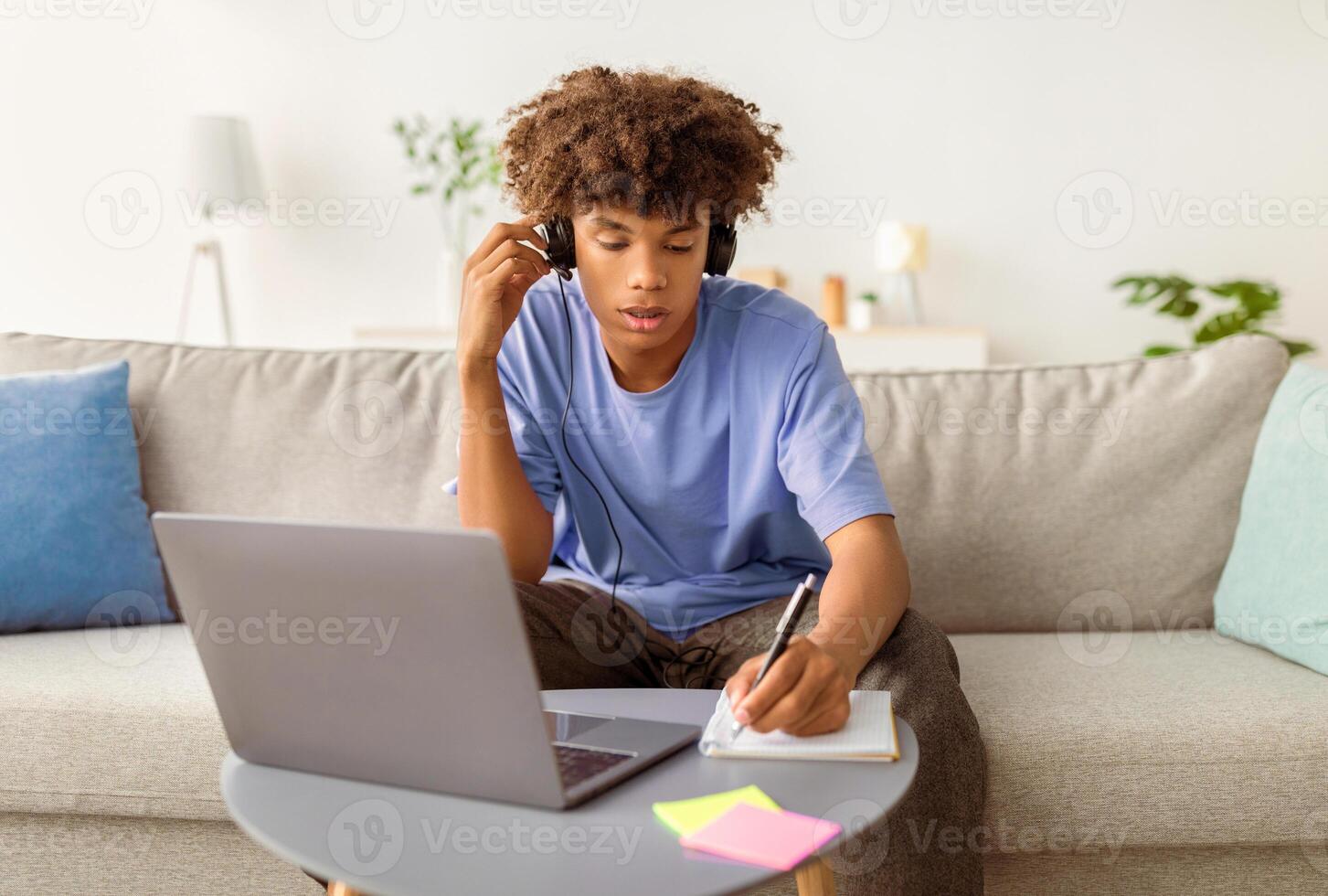 Smart African American teenager attending online lesson or webinar, sitting on couch, using wireless headset and laptop, taking notes indoors. Remote education, home schooling concept photo
