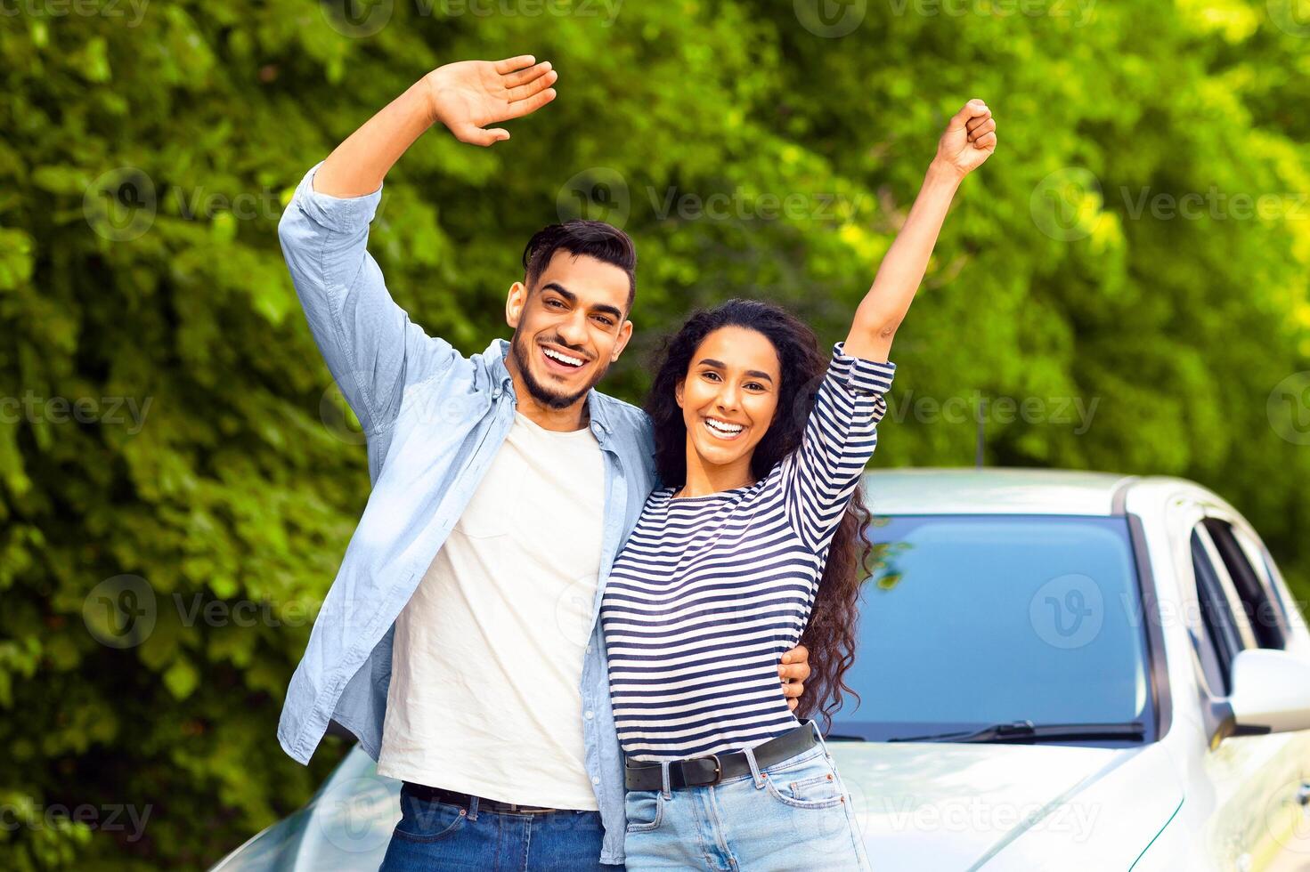 Emotional multiracial couple standing by nice auto at countryside, embracing and waving at camera, copy space. Happy middle-eastern man and woman having car trip together, renting automobile photo