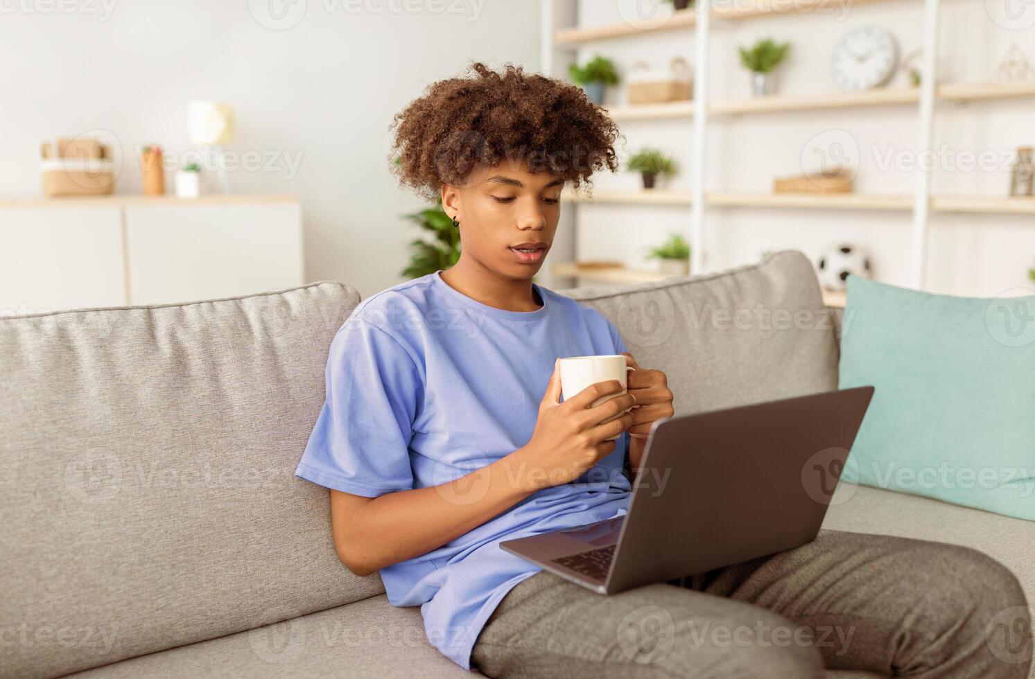 Web-based education. Intelligent black teen boy drinking coffee, having call with fellow students on laptop from home. Afro adolescent participating in online conference photo