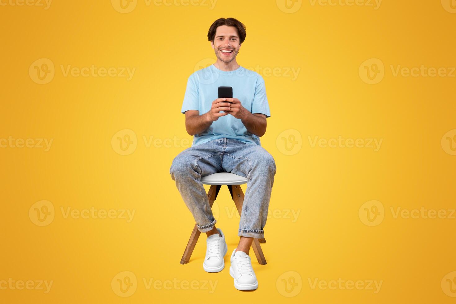People And Technology Concept. Portrait of smiling young man using smartphone sitting on chair isolated on orange studio background, looking at camera. Guy chatting online, browsing social media photo