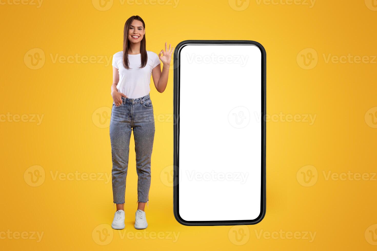 Cool Mobile Offer. Happy casual lady standing near big huge cellphone with empty white screen, showing ok, recommending great new app or website for cell phone, orange wall, mock up, full body length photo