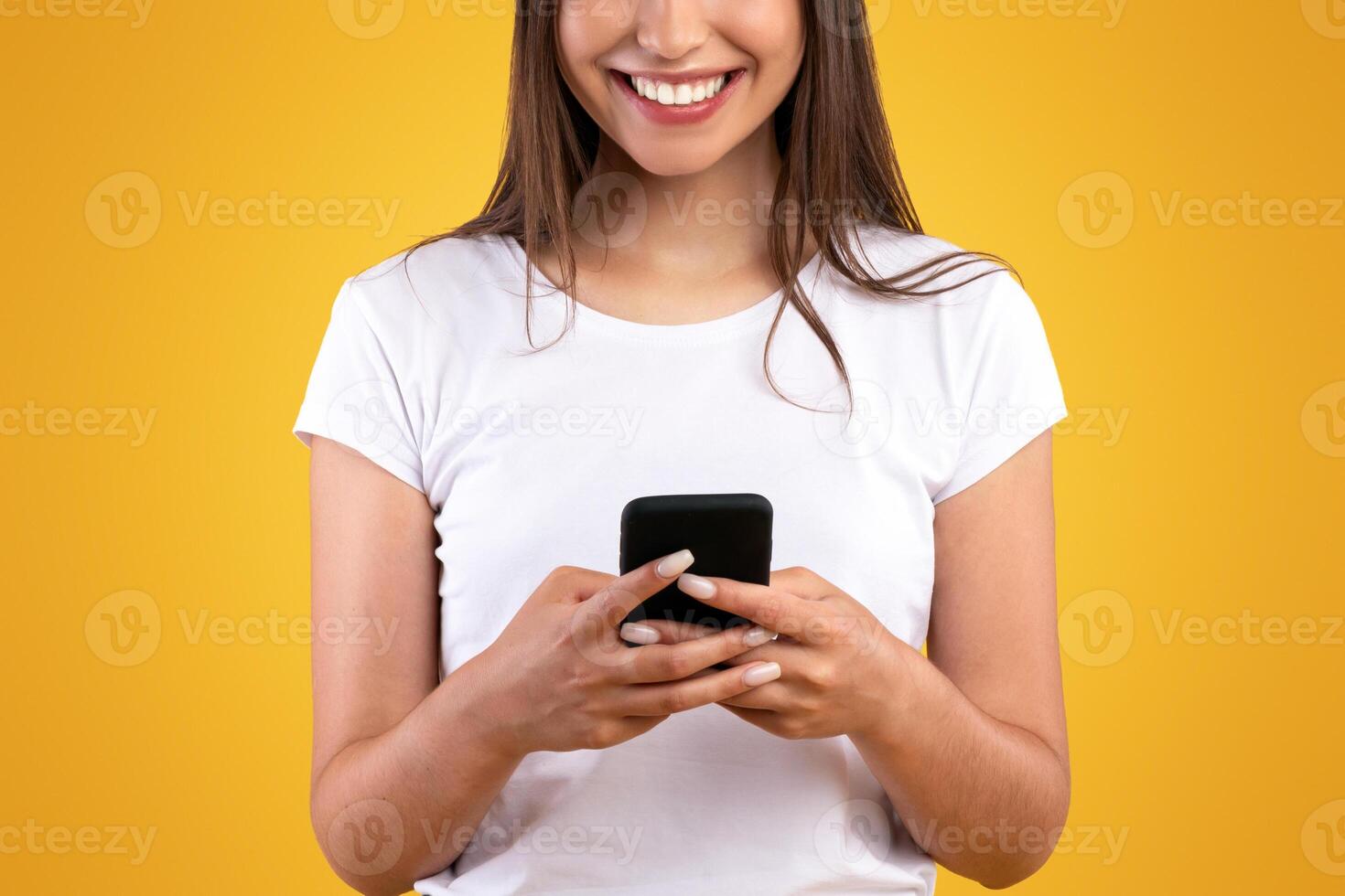 Communication, Technology And Lifestyle Concept. Portrait of smiling young casual lady using mobile phone, standing isolated over orange yellow studio background, chatting, looking at screen photo
