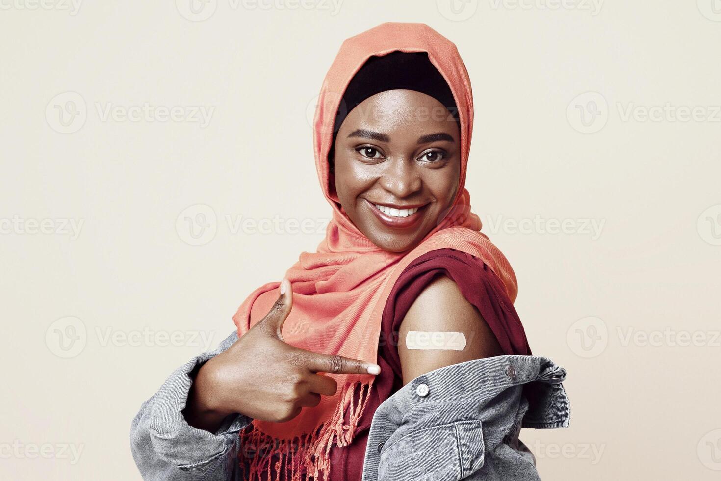 Positive african american millennial muslim woman pointing at adhesive medical band on her shoulder, smiling black lady in hijab got vaccinated against COVID-19, beige studio background, copy space photo