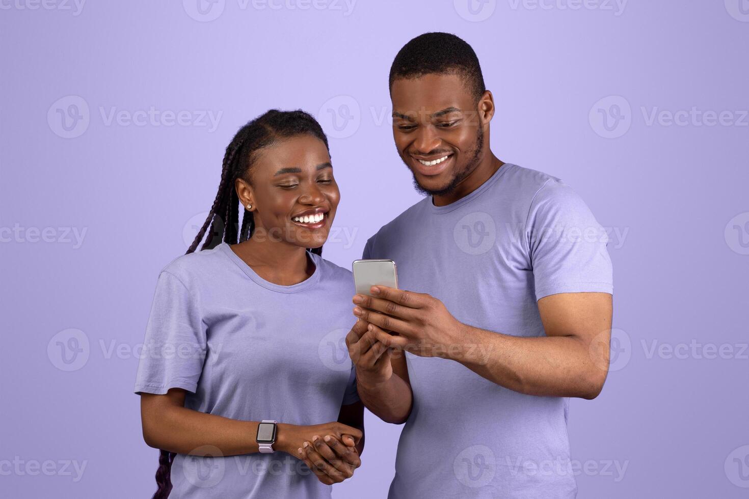 Happy black couple looking at mobile phone screen, using new cool application, violet purple studio background. Joyful African American guy sharing smartphone with lady, modern entertainment concept photo