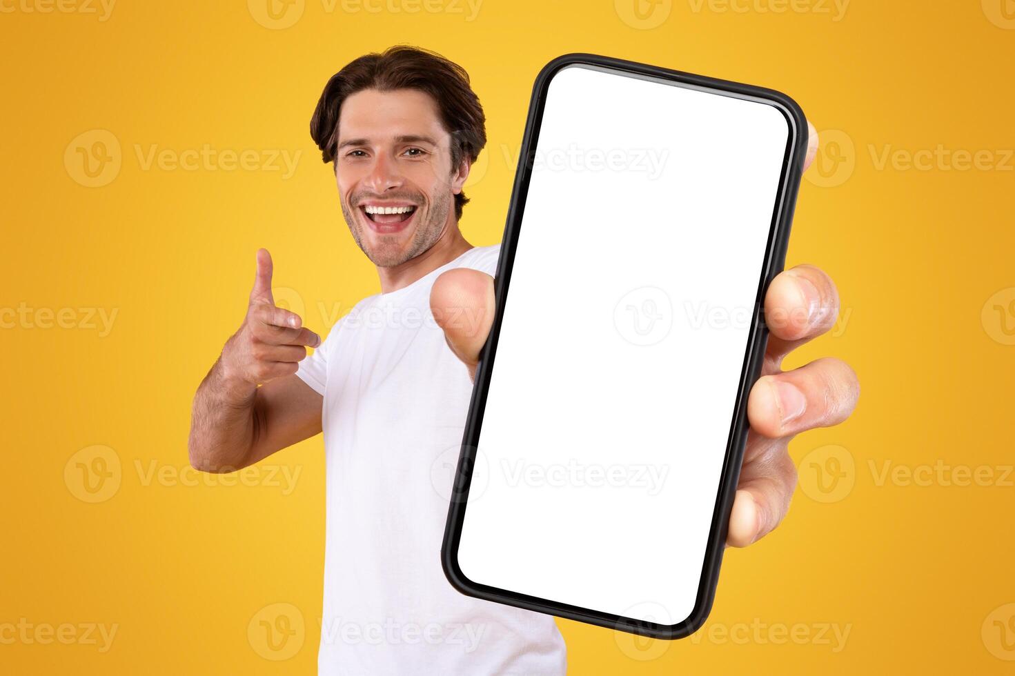 Mobile App Advertisement. Handsome Excited Man Showing Pointing At White Empty Smartphone Screen Posing Over Orange Studio Background, Smiling To Camera. Check This Out, Cellphone Display Mock Up photo