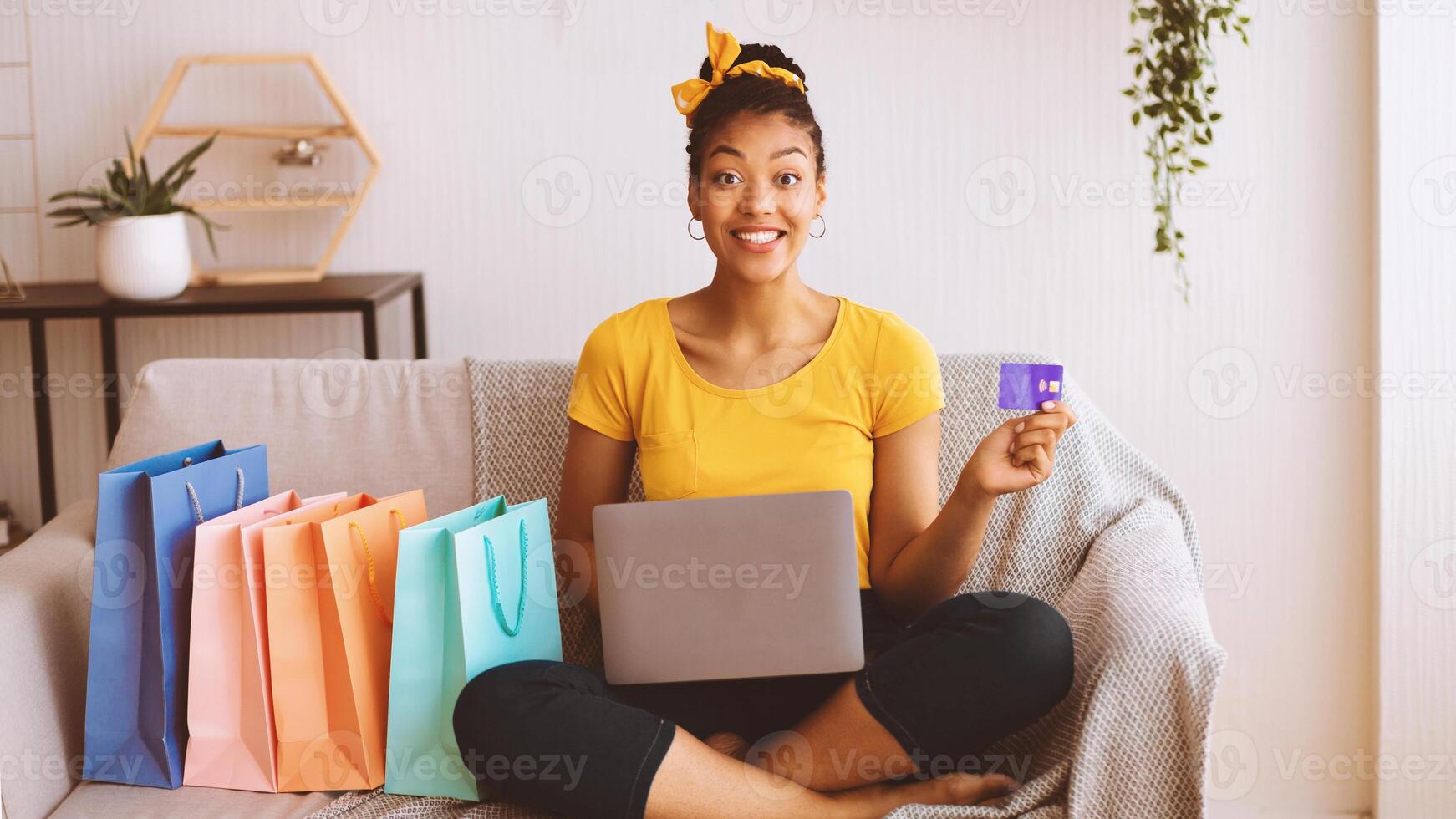 Online Shopping Concept. Excited overjoyed black woman showing credit card and using laptop, ordering clothes and purchases via internet, sitting on the couch with colorful bags in living room at home photo