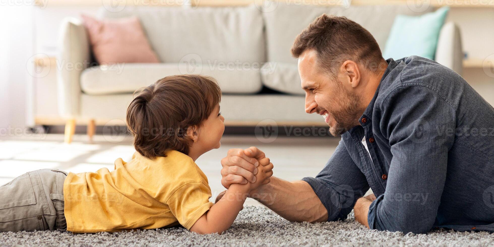 Bringing up strong kid. Happy father competing arm wrestling match with his little boy, lying together on floor at home and smiling to each other, profile portrait photo