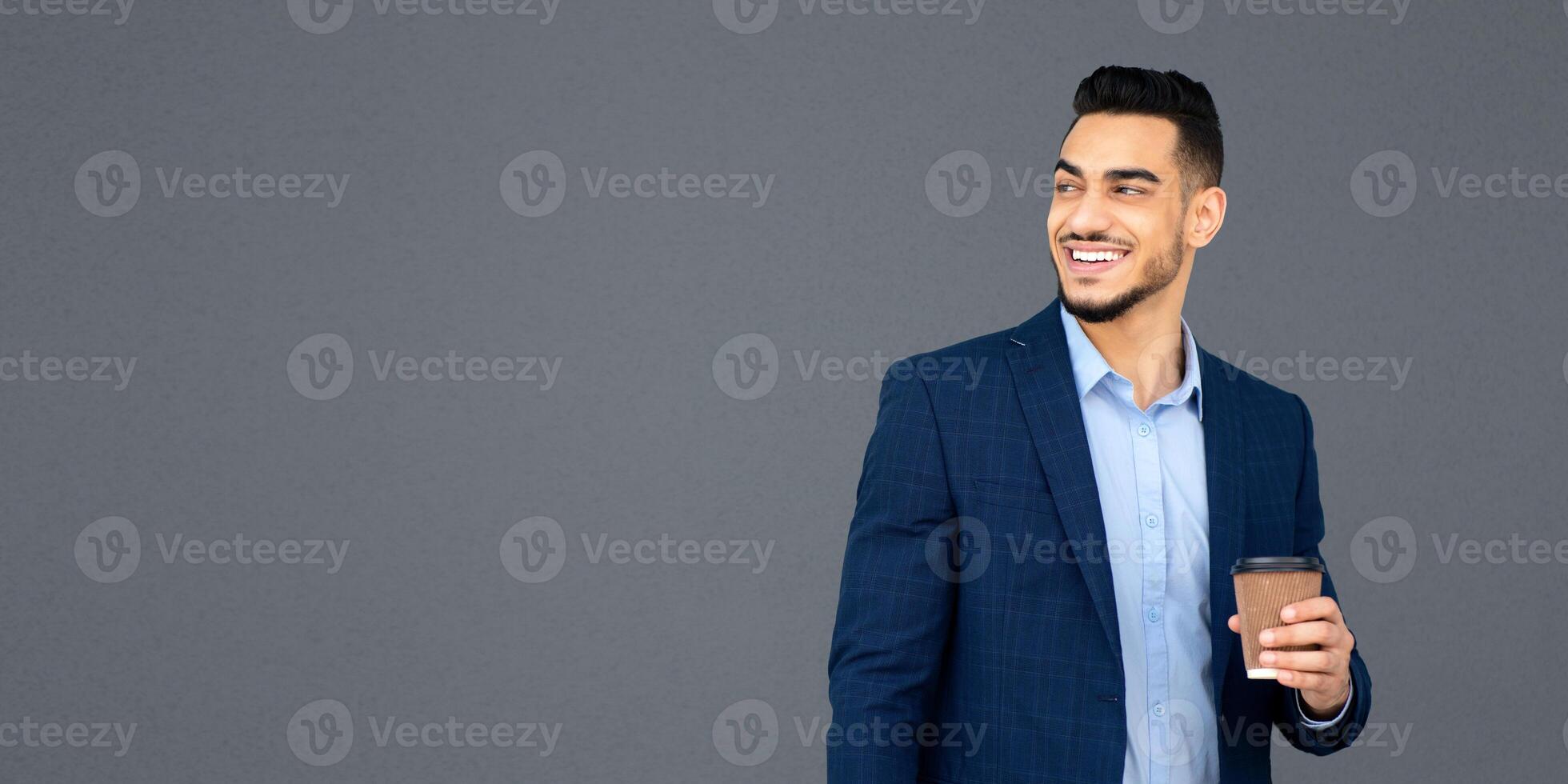 Handsome arab guy businessman in formal outfit with coffee to go and laptop standing outdoors next to grey building wall, looking at copy space, smiling, having break after successful business meeting photo