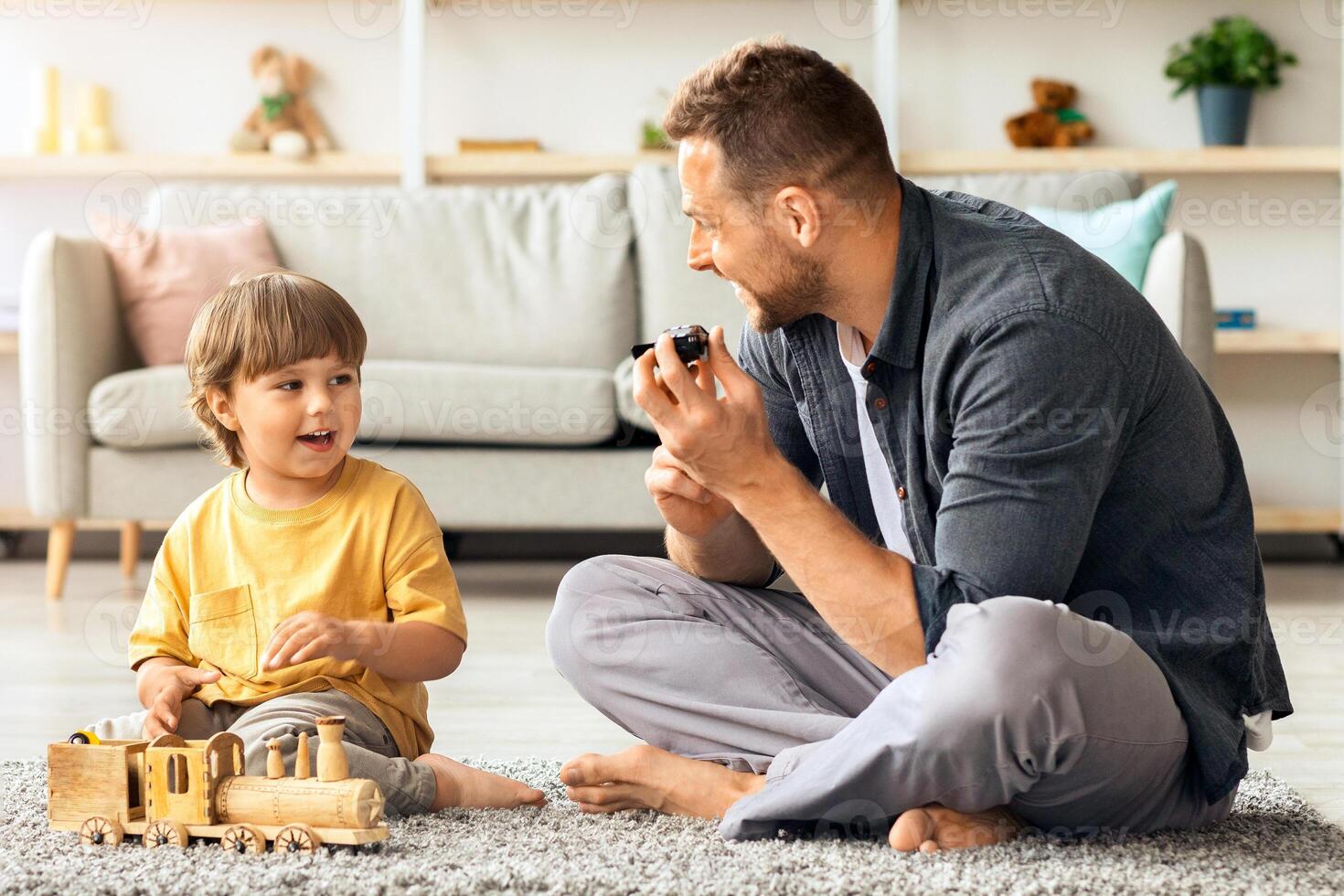 Family pastime. Happy little boy having fun with his daddy at home, playing with toy car and train, sitting on floor at living room, free space photo