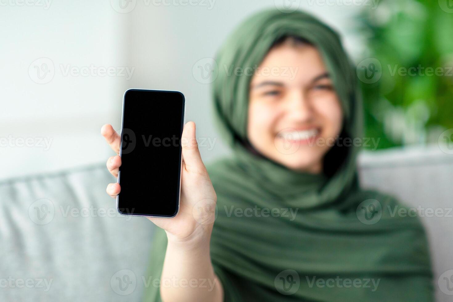 Muslim teenage girl in hijab showing cellphone with blank screen at home, mockup for mobile app or website design. Happy adolescent in headscarf holding smartphone with empty space. Selective focus photo