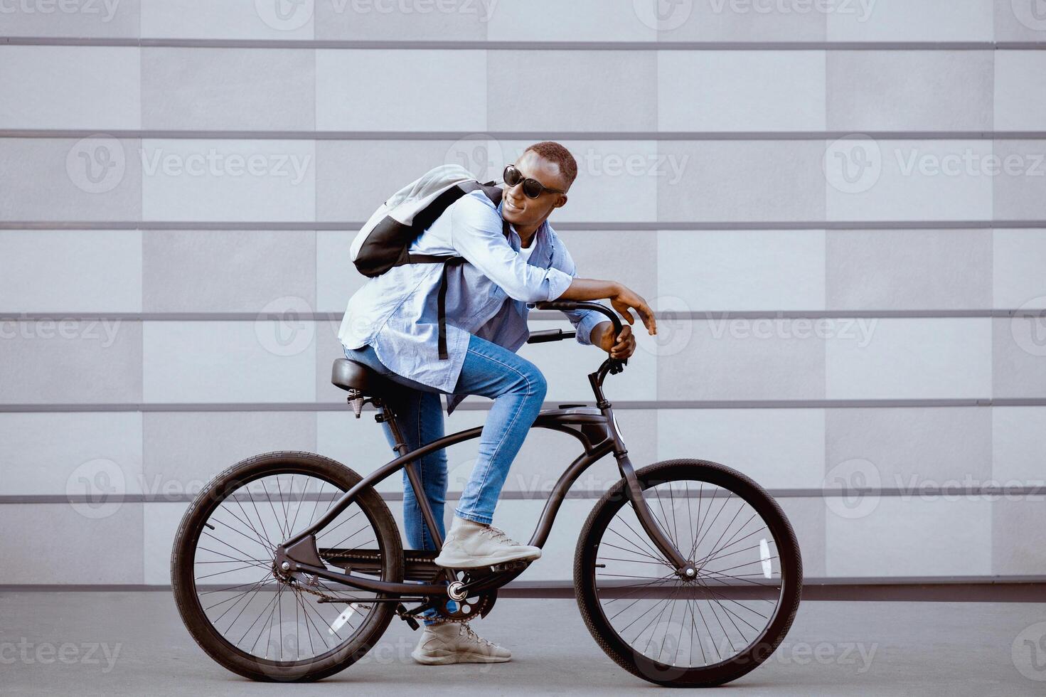 Portrait of handsome black guy with backpack and sunglasses riding bicycle near brick wall outside. Attractive African American hipster traveling in city, going for ride on bike photo