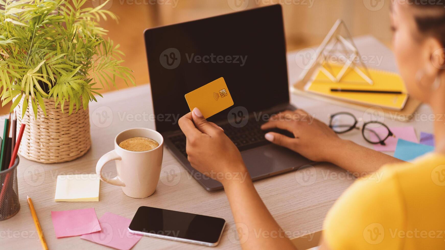 Online Shopping Concept. Over the shoulder view of african american woman holding credit card in hand and using laptop with black blank screen, typing on keyboard, copy space, phone on the table photo